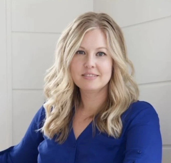 A woman with blonde, wavy hair, wearing a blue top, smiling softly at the camera against a light-colored, paneled background.