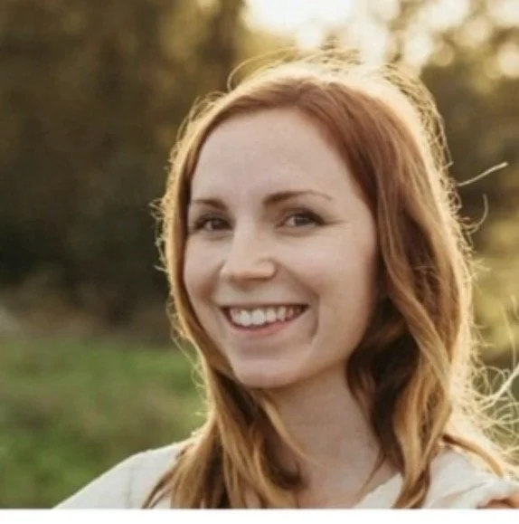 A young woman with red hair smiling outdoors with a blurred background of trees and greenery.