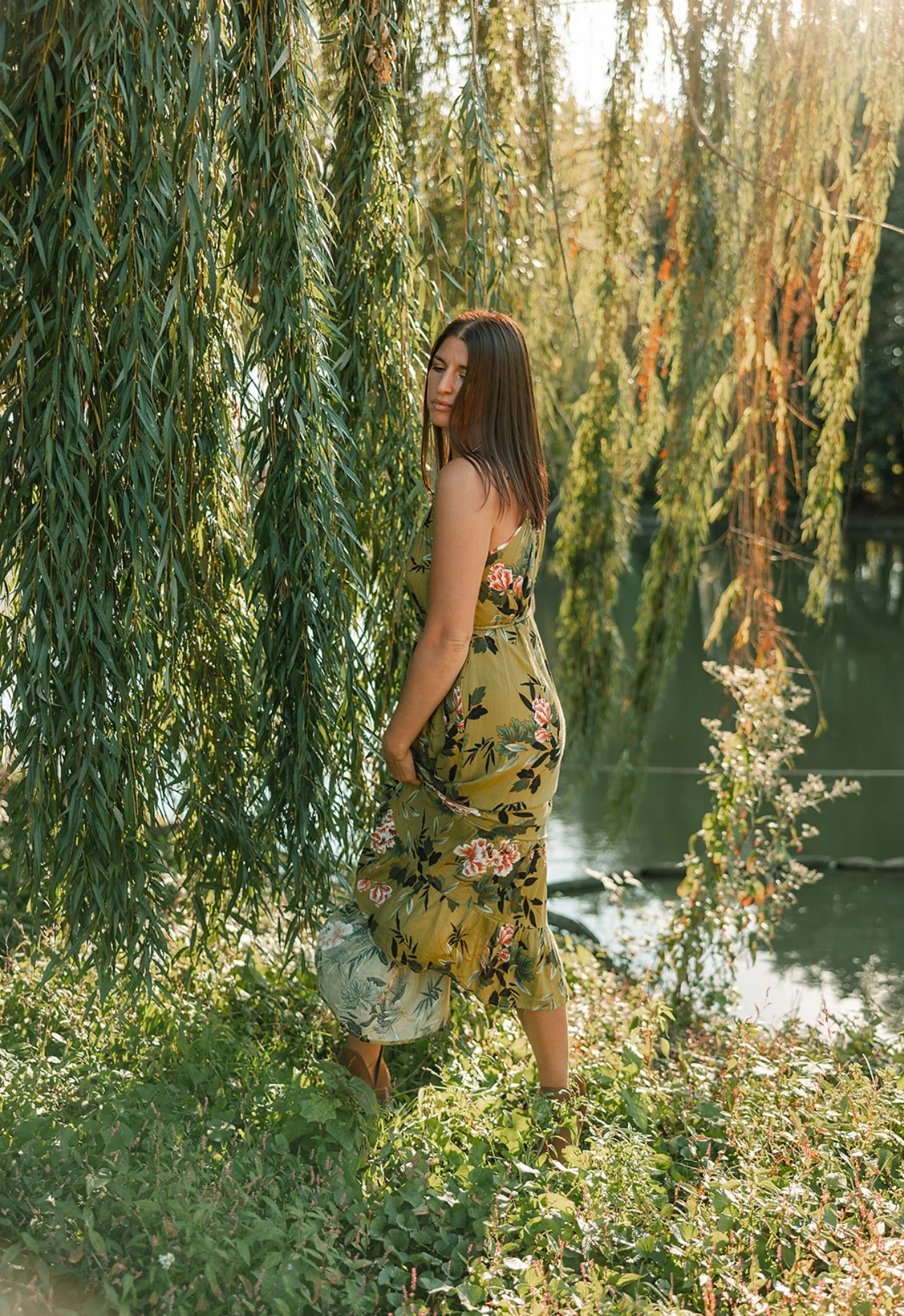 A woman in a floral dress standing near a lush willow tree by a body of water, sunlight filtering through the leaves.