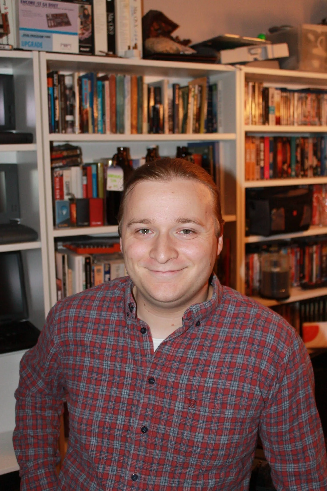 Picture of Kai Griffiths-Meade wearing a red plaid shirt standing in front of white bookshelves filled with books and DVDs.