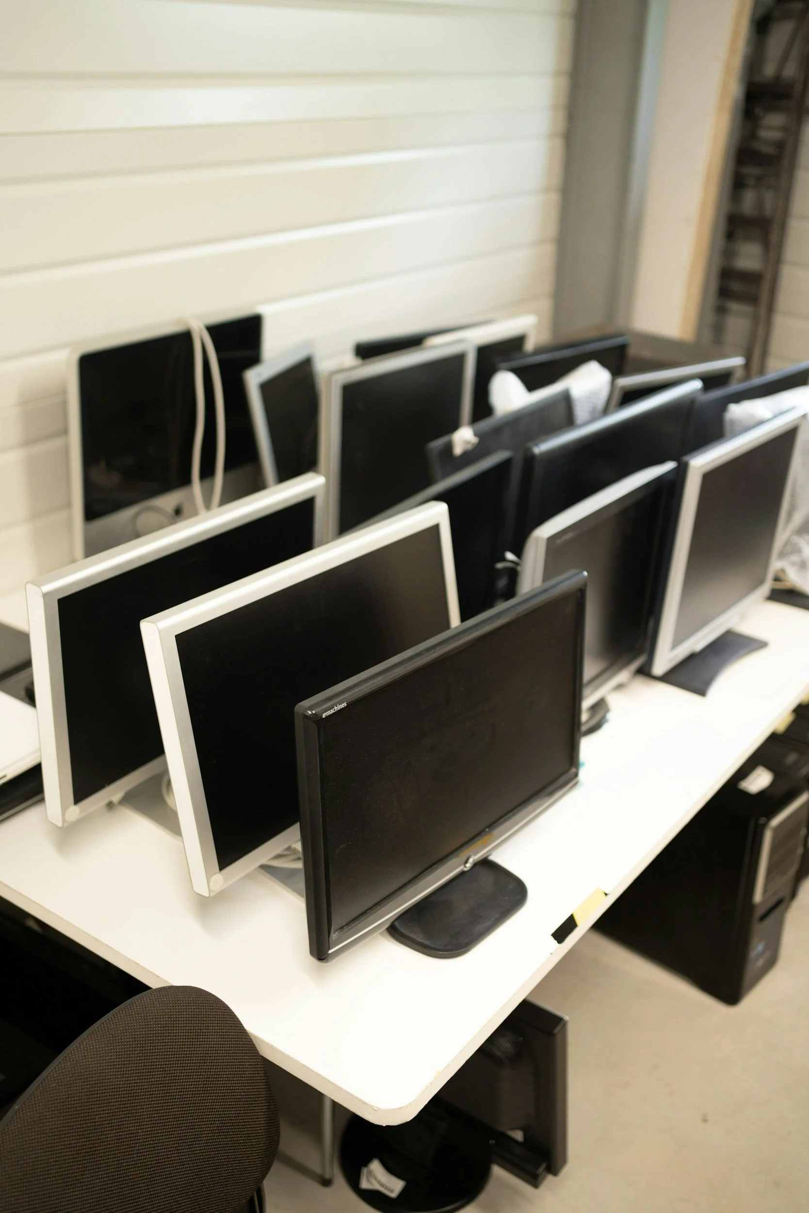 Multiple computer monitors stacked and arranged on a white table in an office or storage room, with some cords hanging and a black desktop tower under the table.