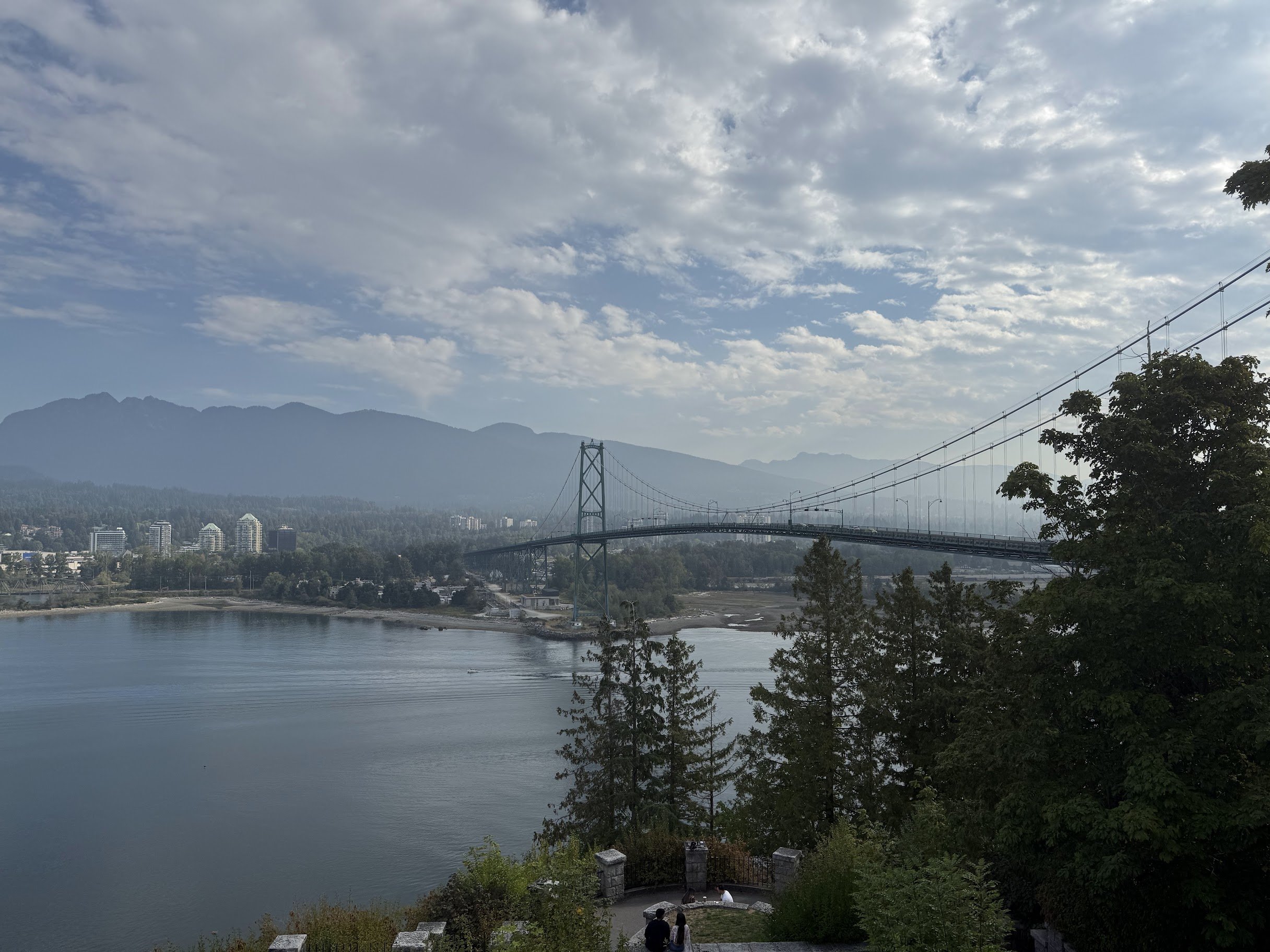 A large suspension bridge over a body of water, with mountains and a partly cloudy sky in the background. Trees and a small viewing area with people in the foreground.