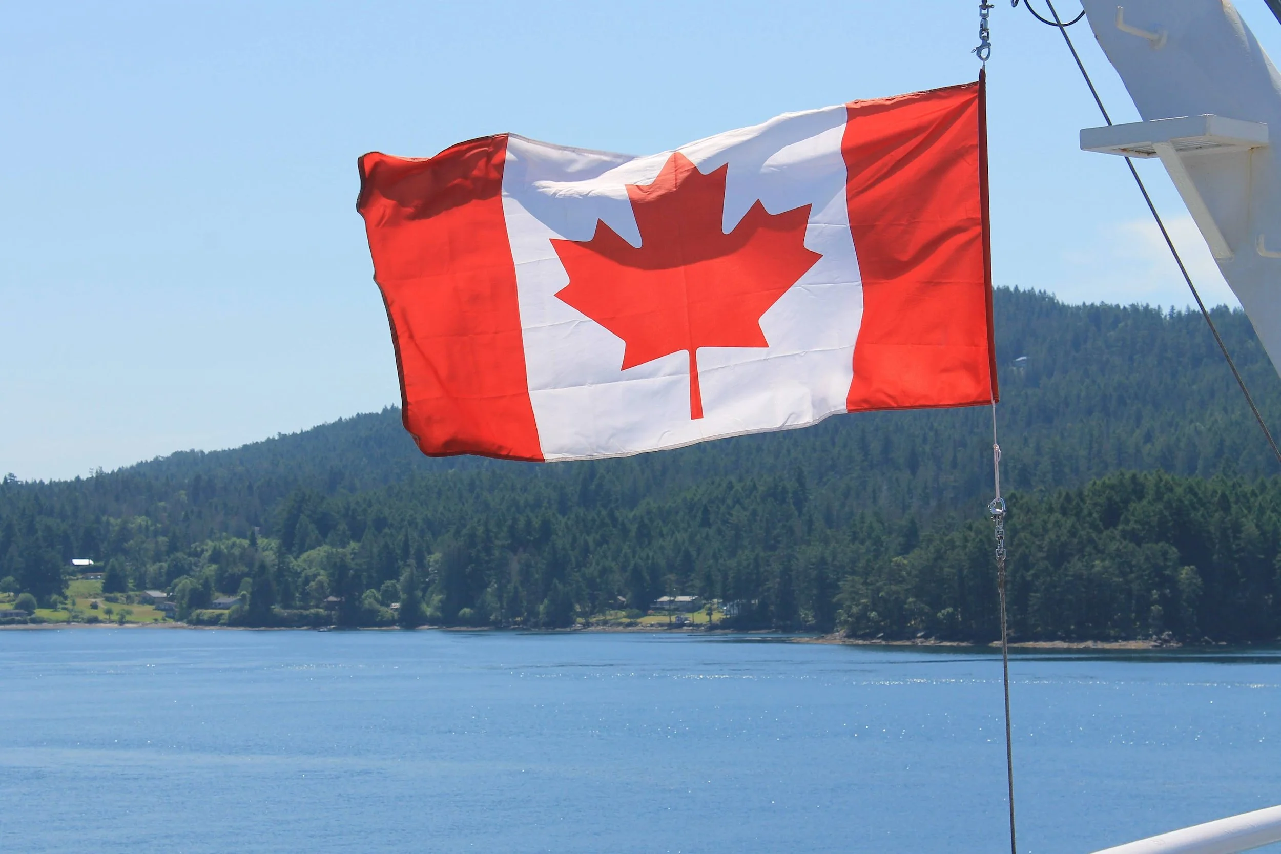 Canadian flag with a red maple leaf on a boat over water with forested hills in the background.