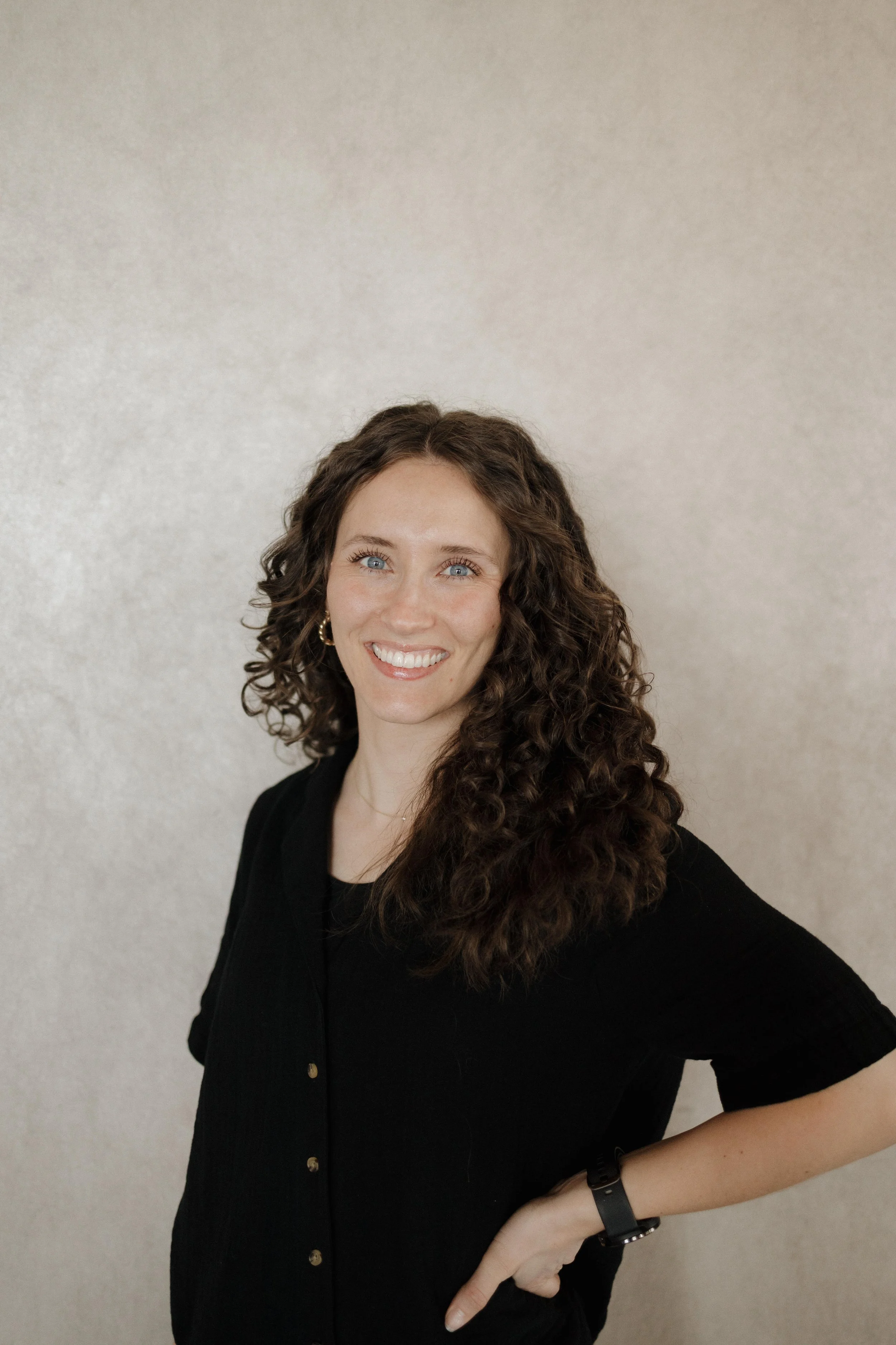 A woman with curly brown hair wearing a black top and gold earrings, smiling against a plain beige wall.