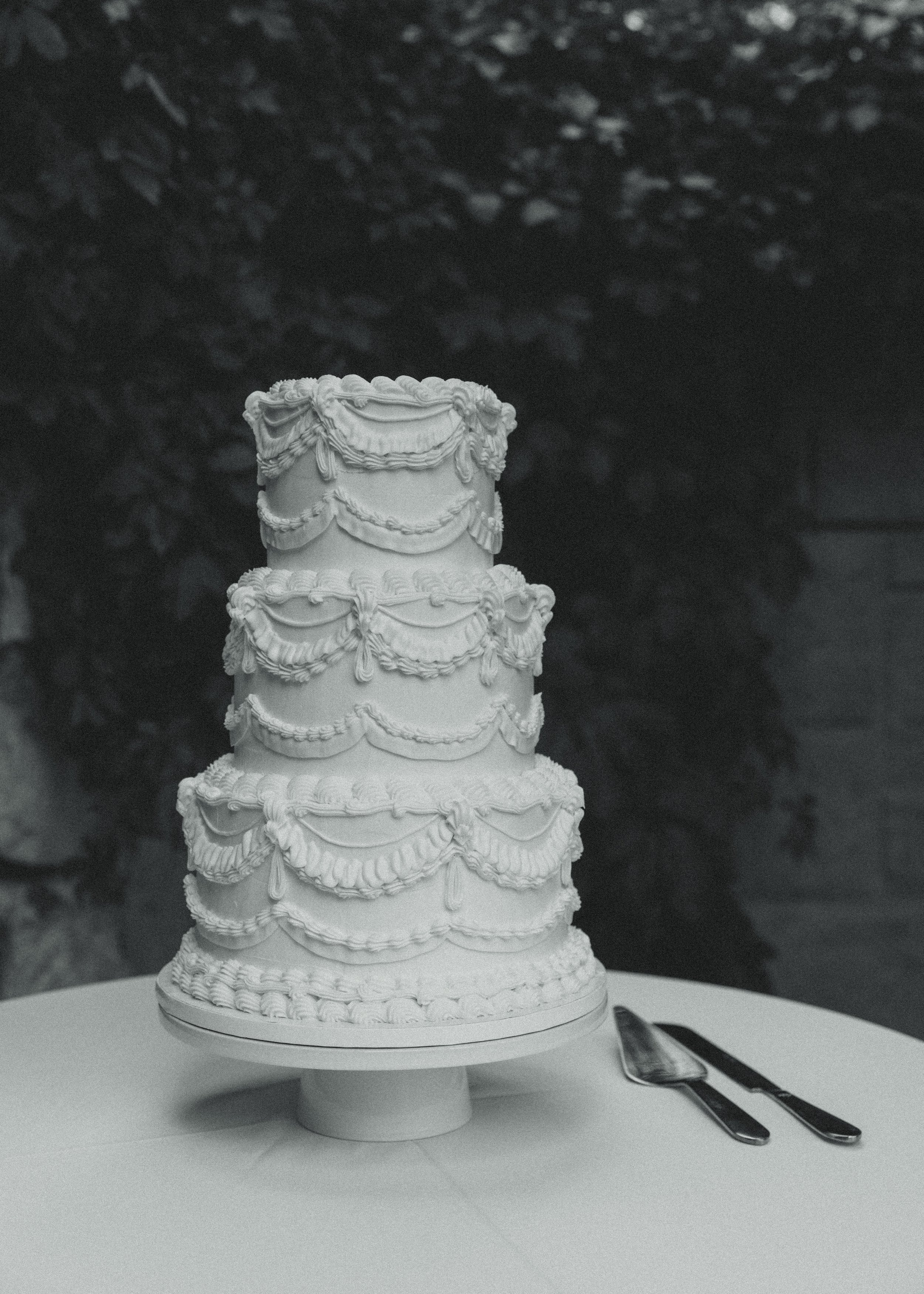 A tall, white, multi-tiered cake with intricate decorative icing, placed on a stand on a round table. Silver cake knife and server are beside the cake. The background shows some trees and a wooden fence.