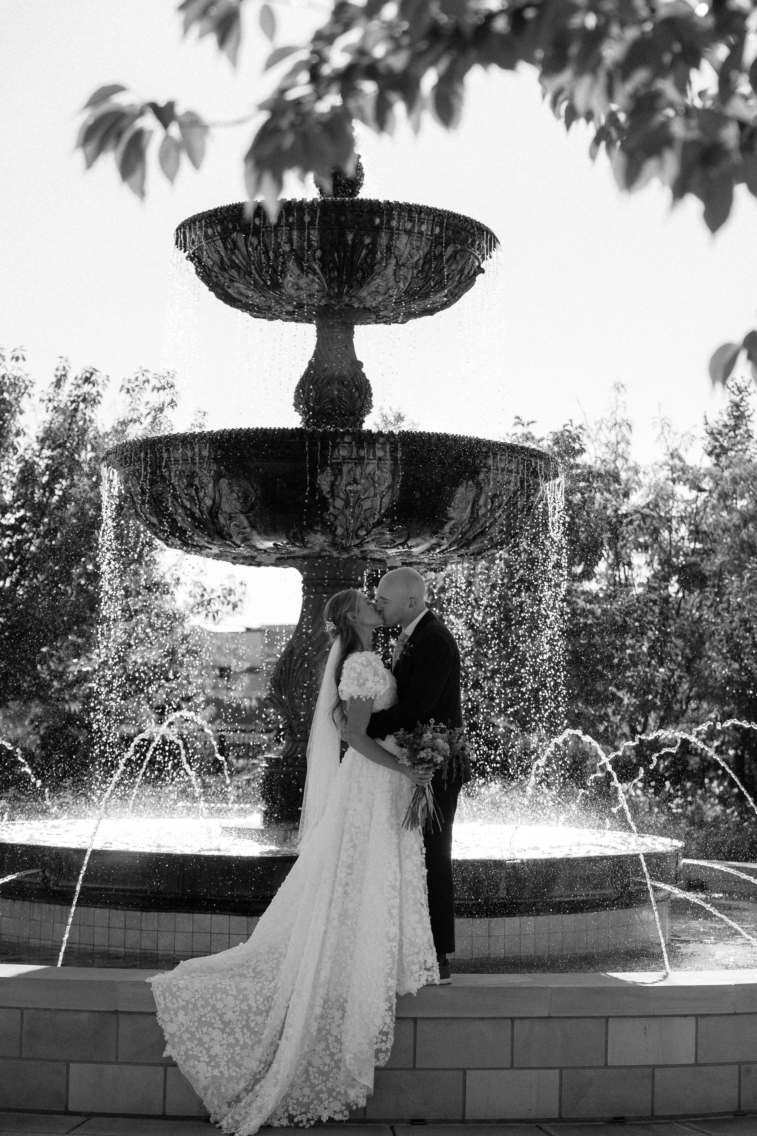A bride and groom sharing a kiss in front of a large ornate fountain with water cascading down, trees and foliage in the background.