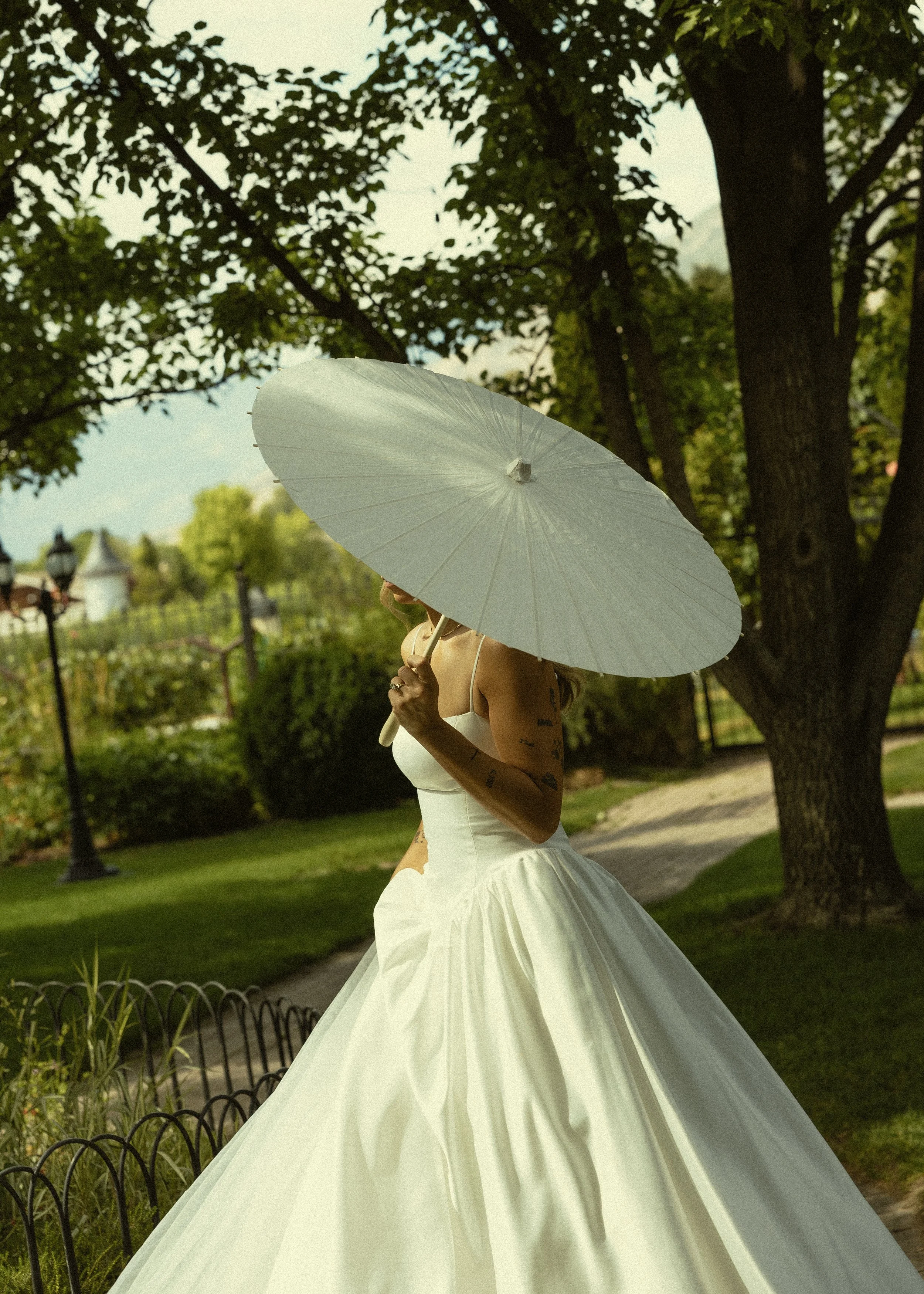 A person wearing a white dress holding a white parasol outdoors in a garden, with trees and a pathway in the background.