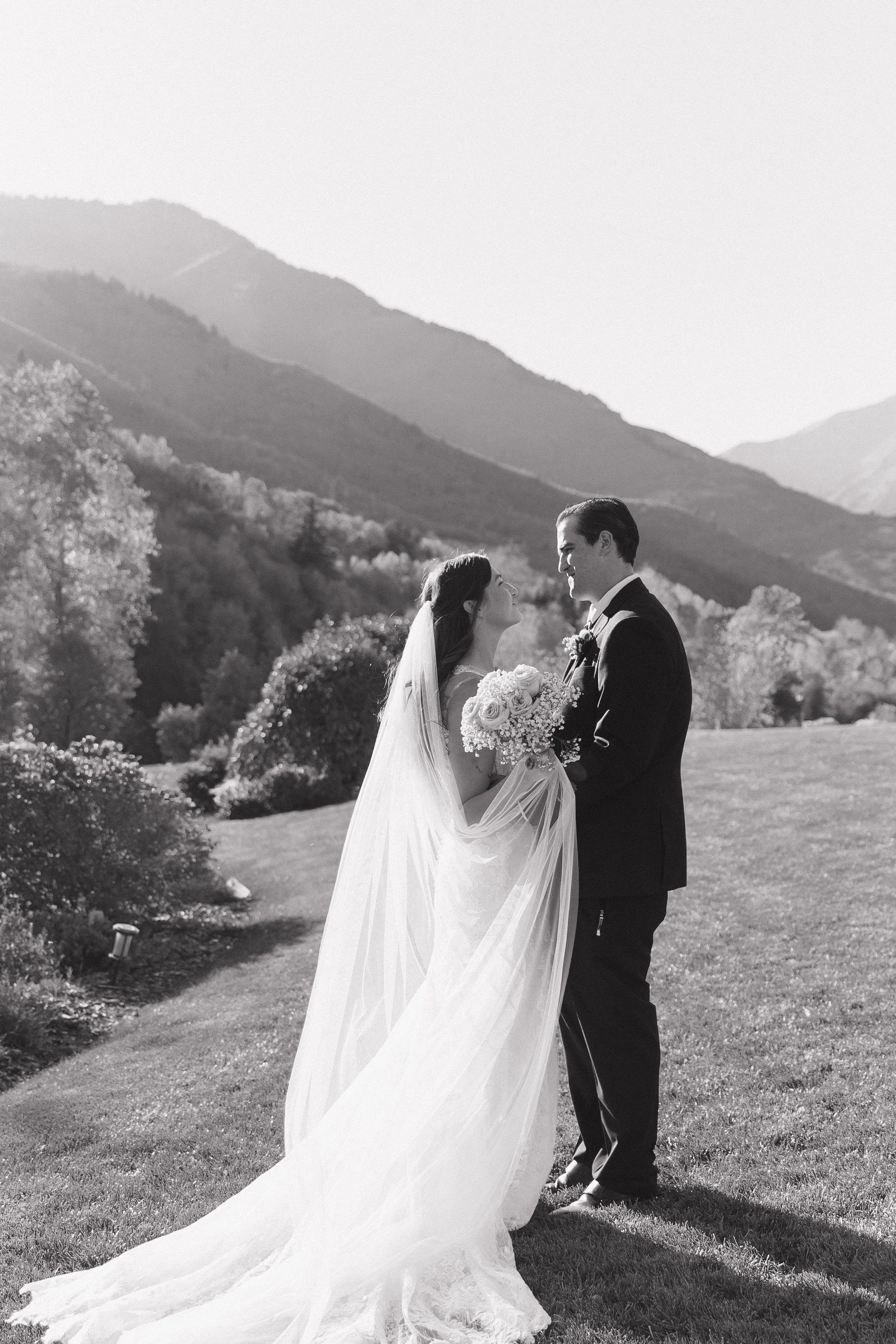 A black and white photo of a bride and groom standing outdoors on a grassy hill, holding a bouquet of flowers, with mountains and trees in the background.