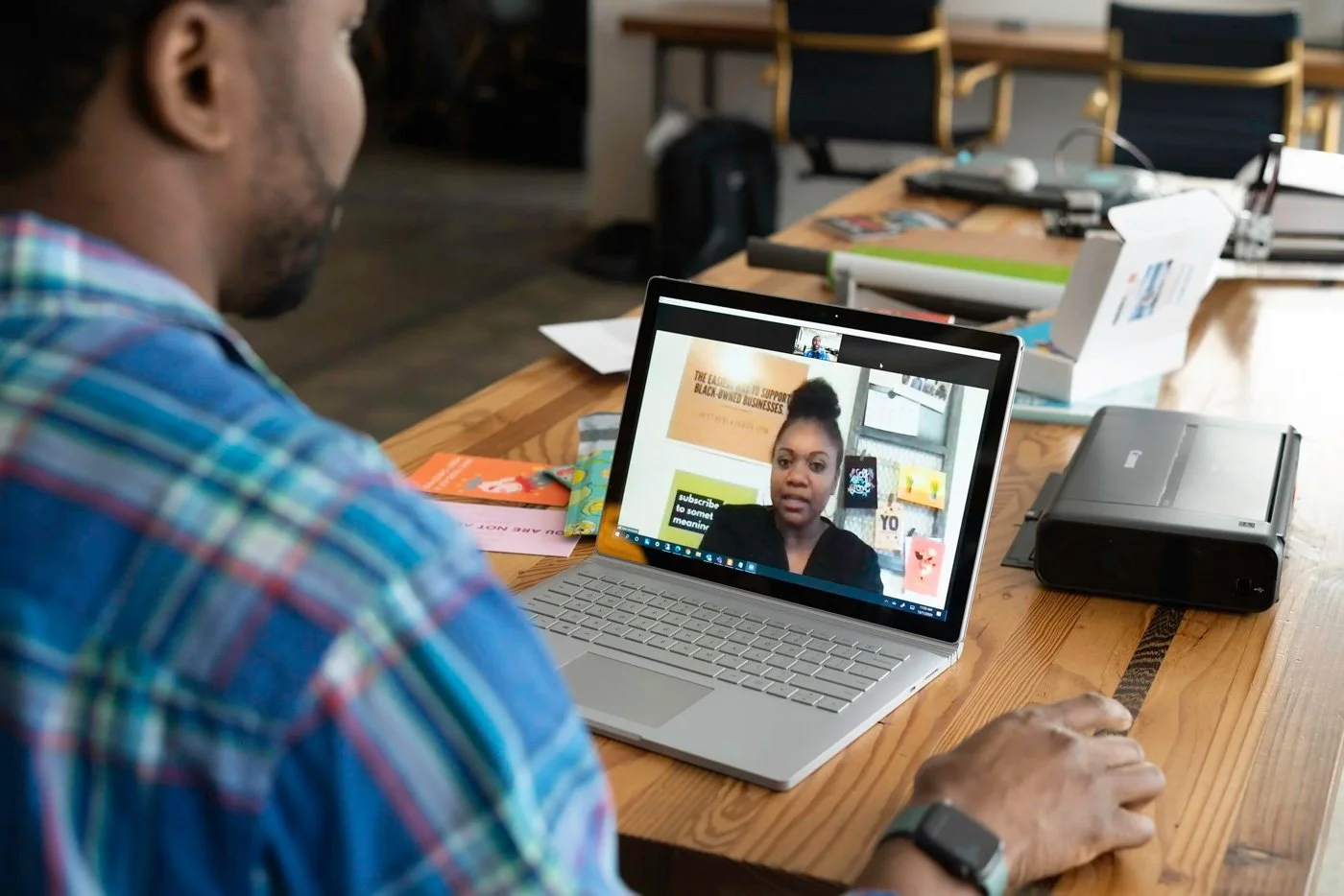 A man sitting at a wooden table engaging in a video call on a laptop, with papers, a printer, and office supplies on the table.