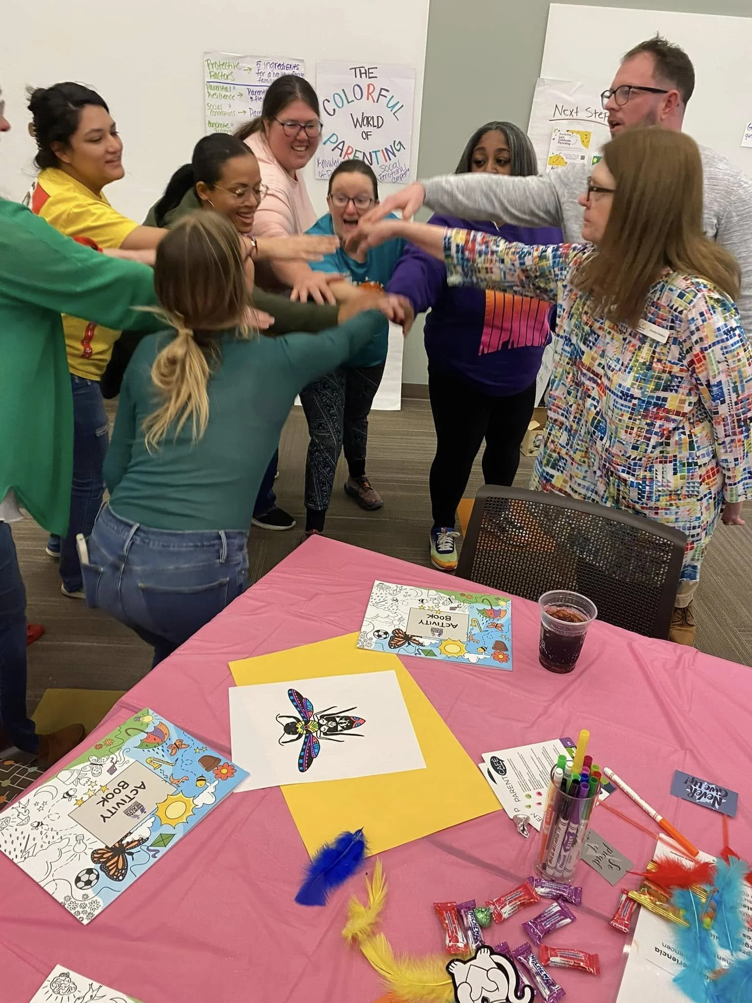 A group of people standing around a table with coloring pages, markers, and feathers, engaging in a group activity or game in a room with colorful posters.