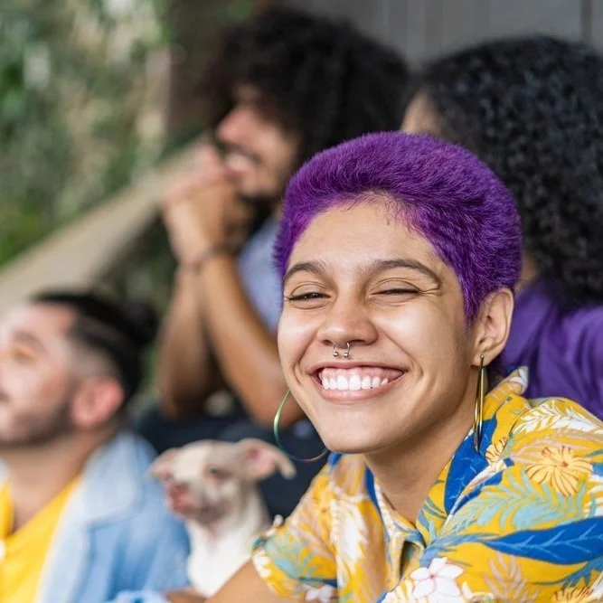 Group of friends smiling outdoors, one with purple hair, wearing a colorful Hawaiian shirt, with a dog in front of them.