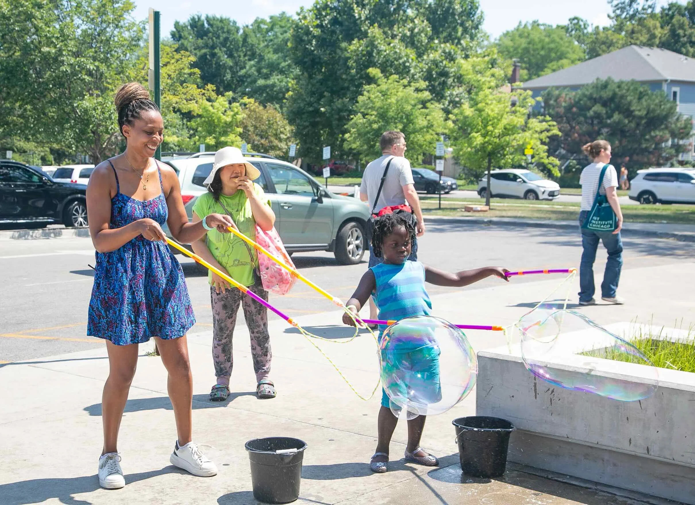 A woman and children blowing soap bubbles outdoors on a sunny day in a parking lot, with trees and buildings in the background.