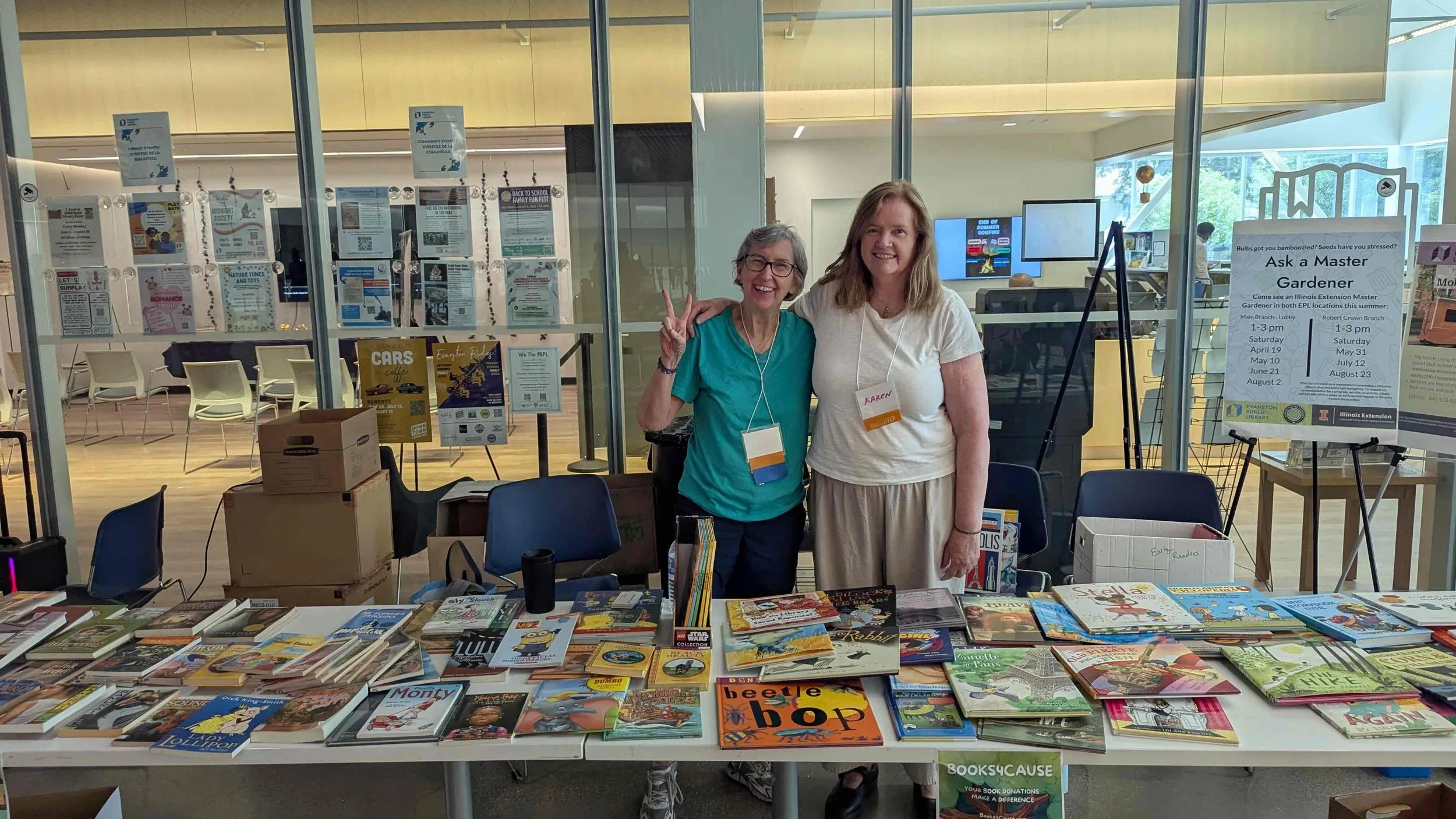 Two women stand behind a table filled with books and promotional materials inside a library. They are smiling and making peace signs, wearing name tags, with a glass wall and additional desks in the background.