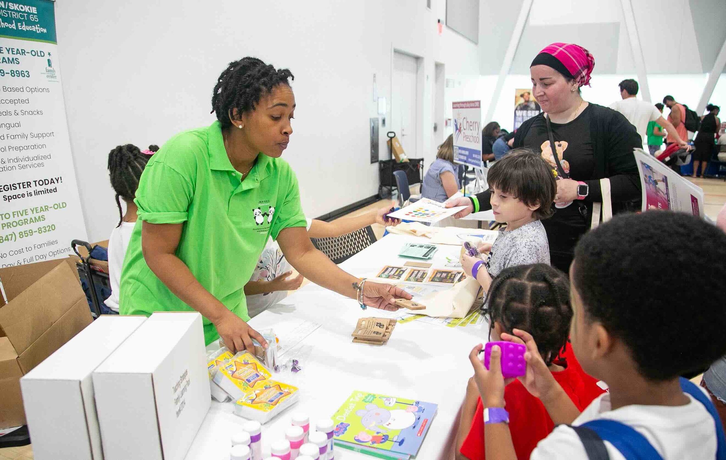 A woman in a bright green shirt handing out items to children at a booth inside a busy event space.