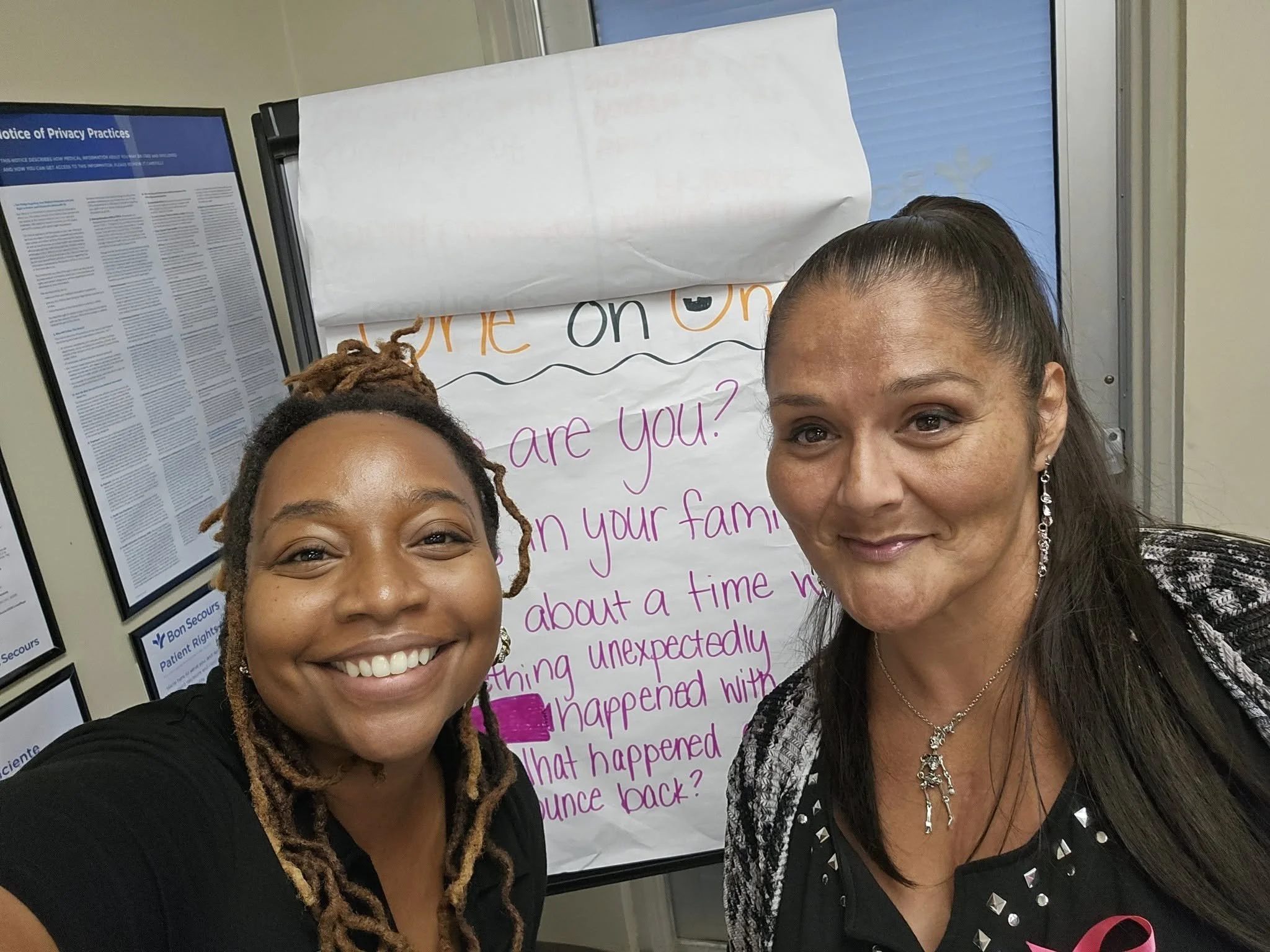 Two women are smiling for a selfie in front of a handwritten poster and informational posters on the wall. One woman has long dreadlocks, and the other has long straight hair. The poster behind them has purple and black writing on white paper, with some visible words like "about a time" and "happened." The informational posters include text and are framed.