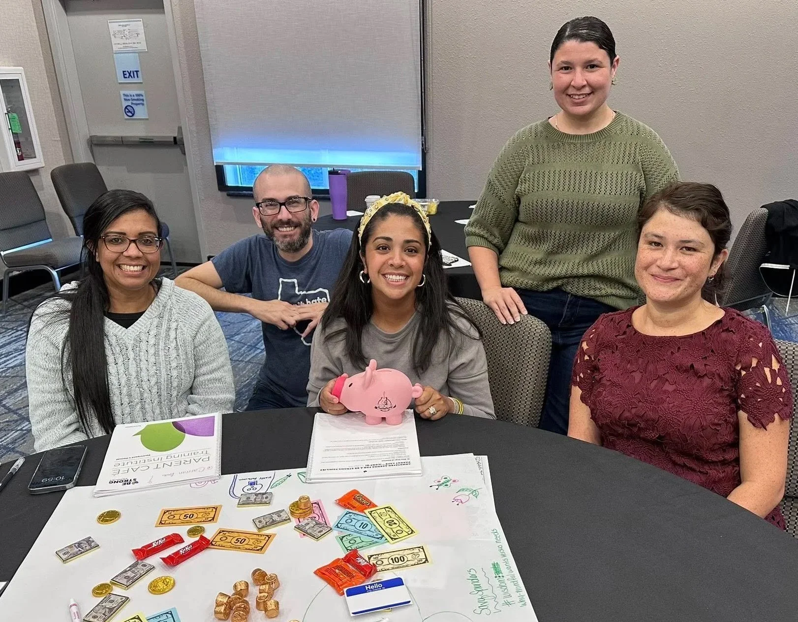 A group of five smiling people gathered around a table with play money, snacks, and a pink piggy bank, in a conference room.
