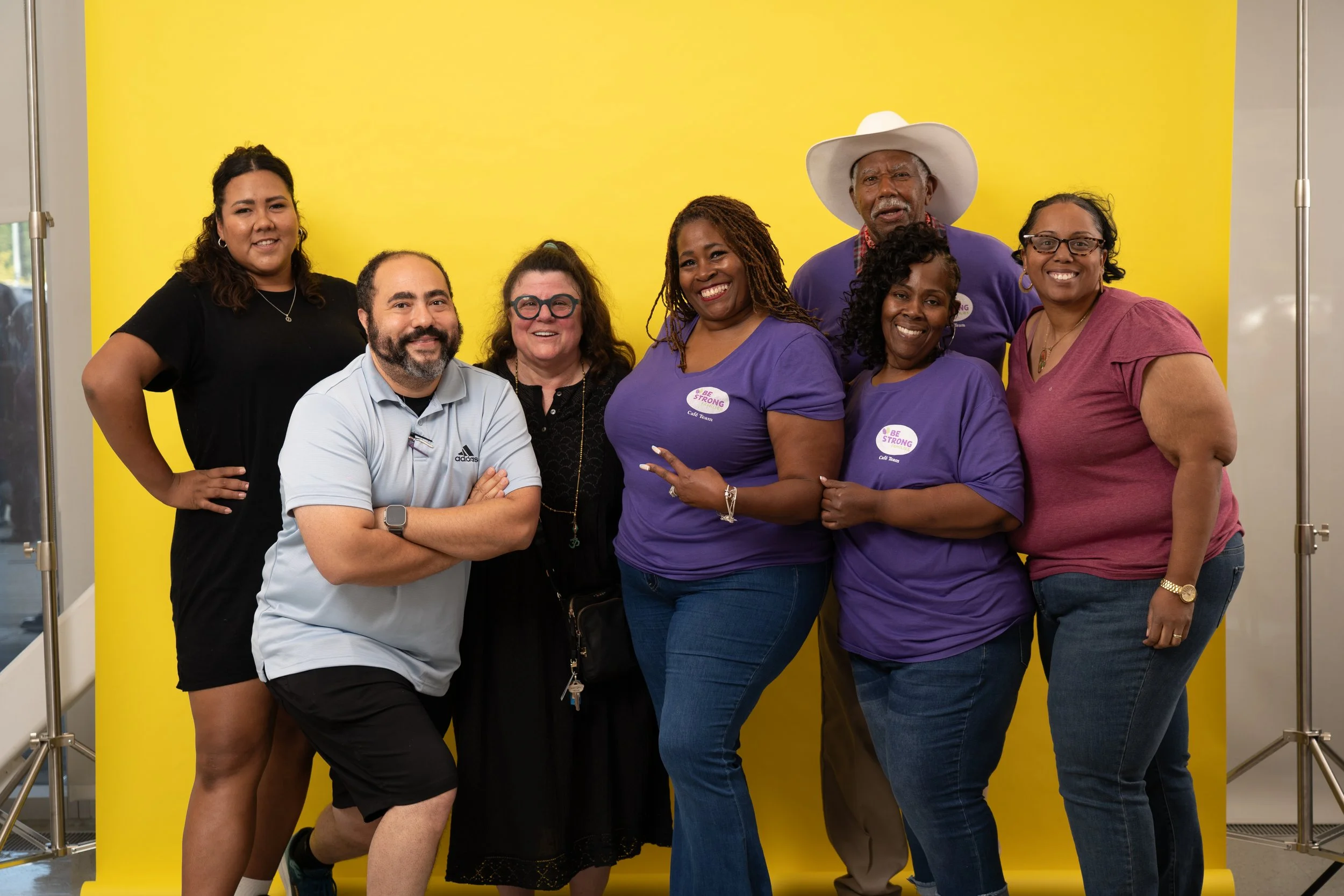 A group of eight diverse smiling people standing in front of a yellow backdrop indoors. Some are wearing purple t-shirts with a 'Be Strong' sticker, and one person is wearing a cowboy hat. They appear happy and are posing for a group photo.
