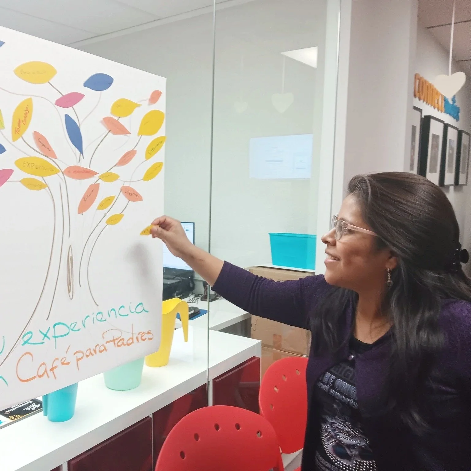 Woman with glasses smiling and inserting a yellow sticky note on a colorful poster calendar in an office.
