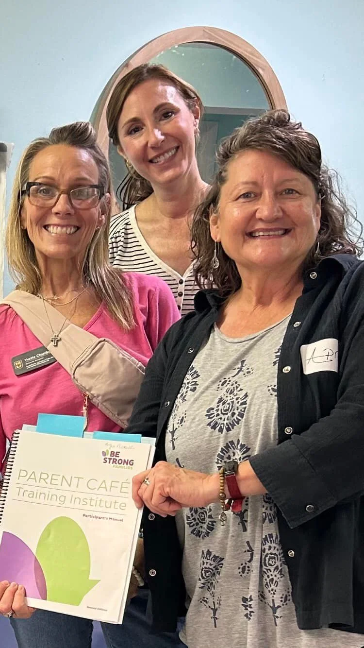 Three women smiling at a training event, with one holding a booklet titled 'Parent Cafe Training Institute'.