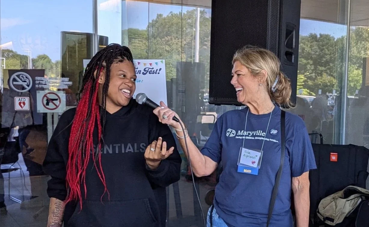 Two women are smiling and laughing as one holds a microphone toward the other during an outdoor event. The woman on the left has long, braided hair with red tips and wears a black hoodie. The woman on the right has blonde hair tied back, wears a blue t-shirt with a name tag, and has headphones around her neck.
