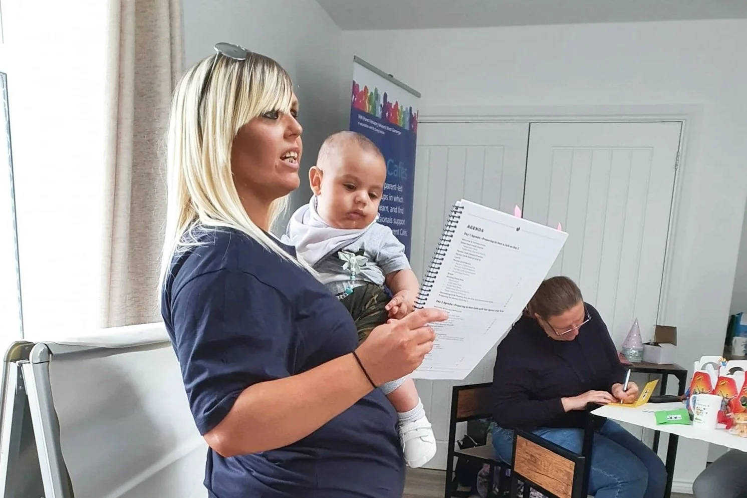 Woman holding an infant and reading from a guidebook to a room of people