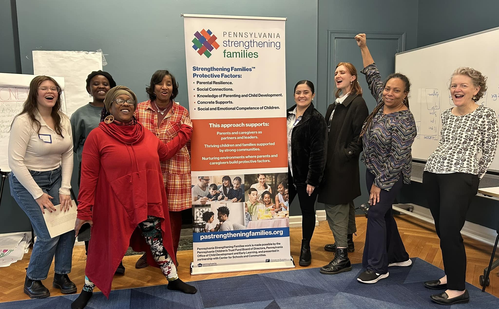 A diverse group of women standing and smiling next to a banner for Pennsylvania strengthening families. The women are in a room with blue walls, whiteboards, and various papers. One woman is raising her fist in celebration.