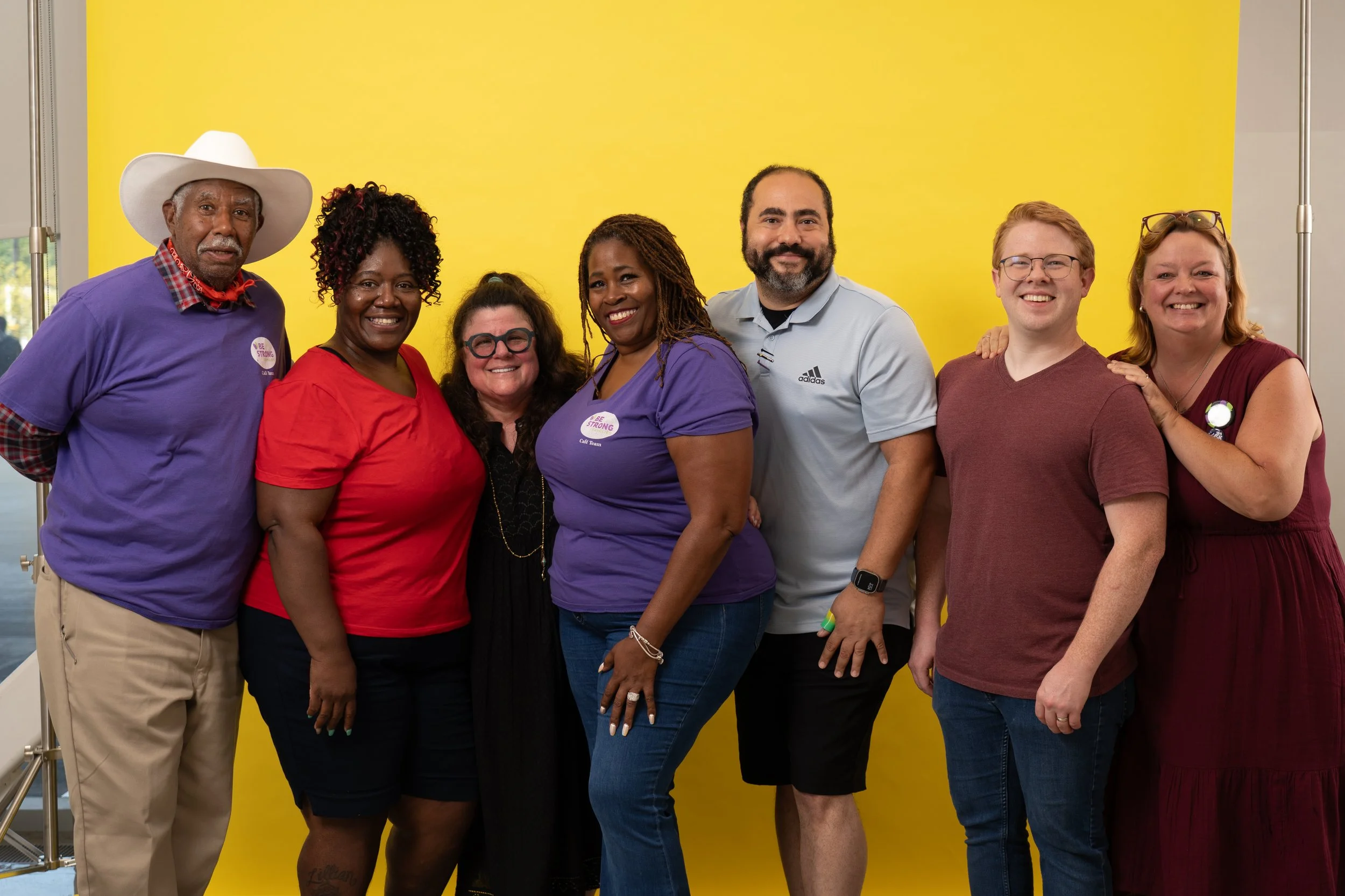 Group of seven diverse adults standing in front of a bright yellow background, smiling for the camera.