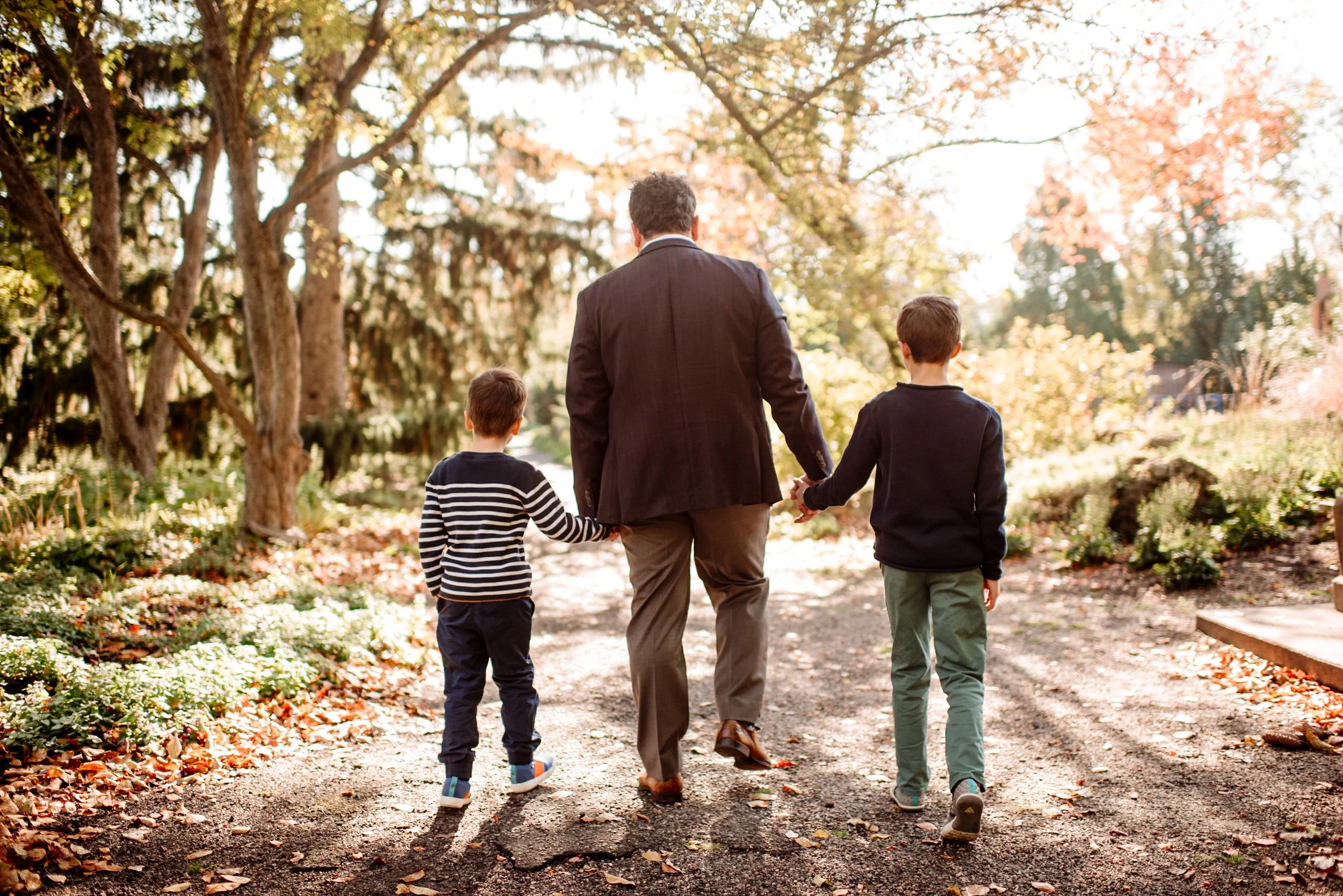 A man in a dark suit walking hand-in-hand with two boys along a leaf-strewn trail in a park during autumn.