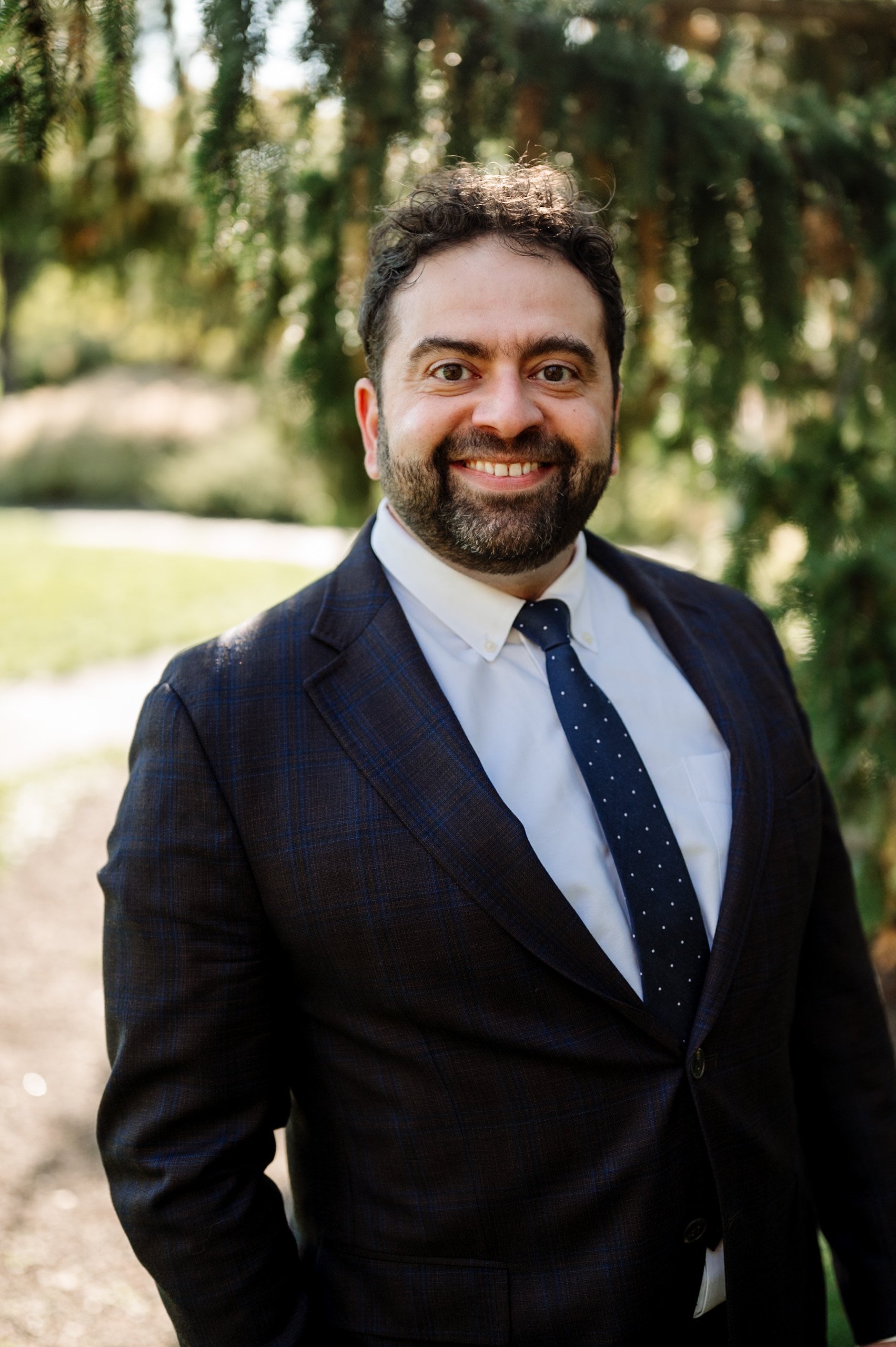 A man with dark, curly hair and a beard, smiling, wearing a dark suit, white shirt, and navy blue tie with white dots, outdoors with green trees and blurred background.