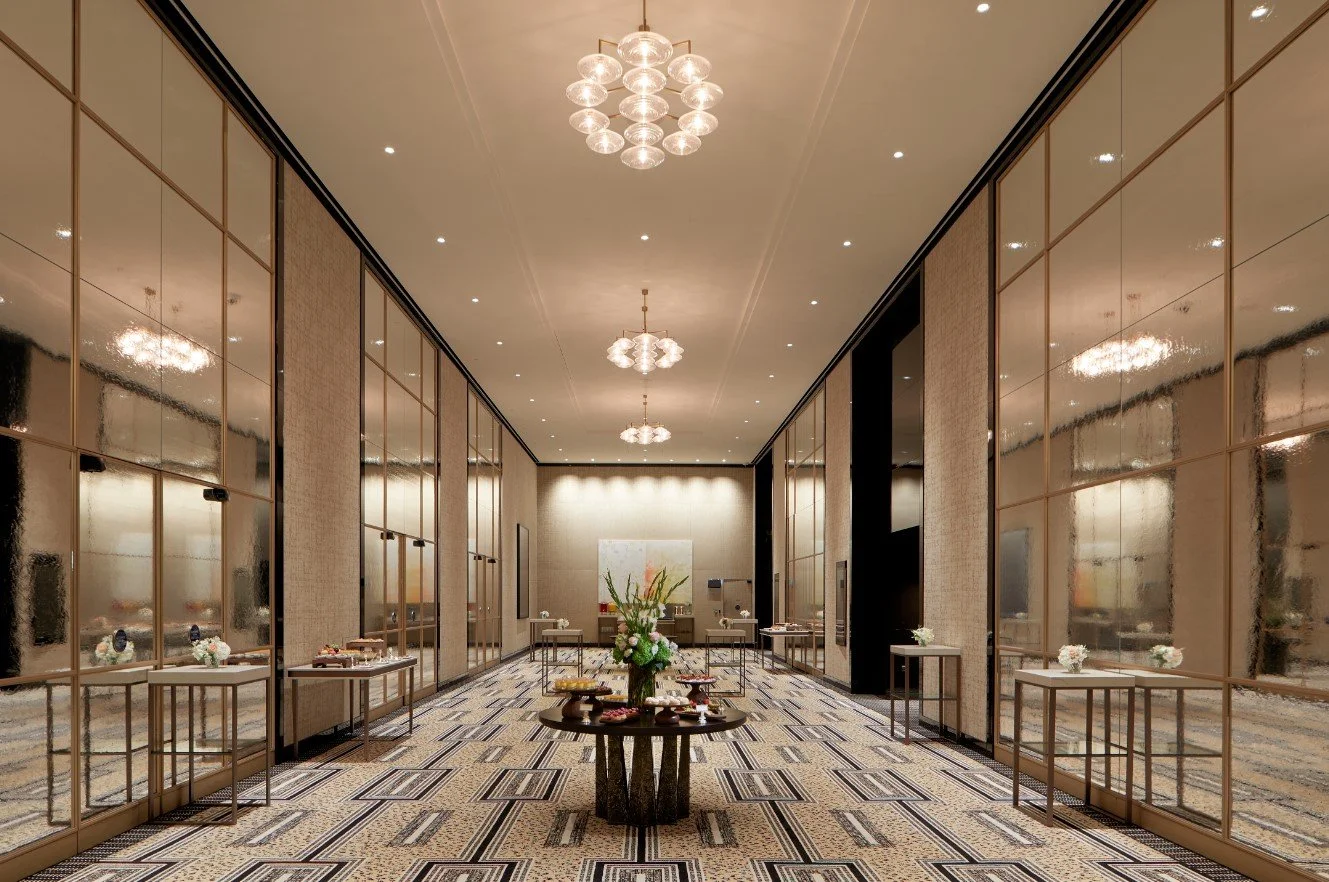 Elegant hotel lobby with a high ceiling and modern chandeliers, featuring mirrored wall panels, a patterned carpet, and tables with flower arrangements.