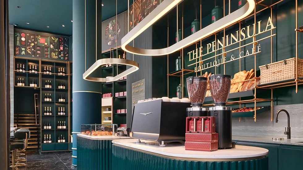 Interior of a stylish cafe named 'The Peninsula Boutique & Café' with a modern design, featuring dark green walls, gold accents, a coffee counter with two coffee grinders, and shelves with baked goods and beverages.