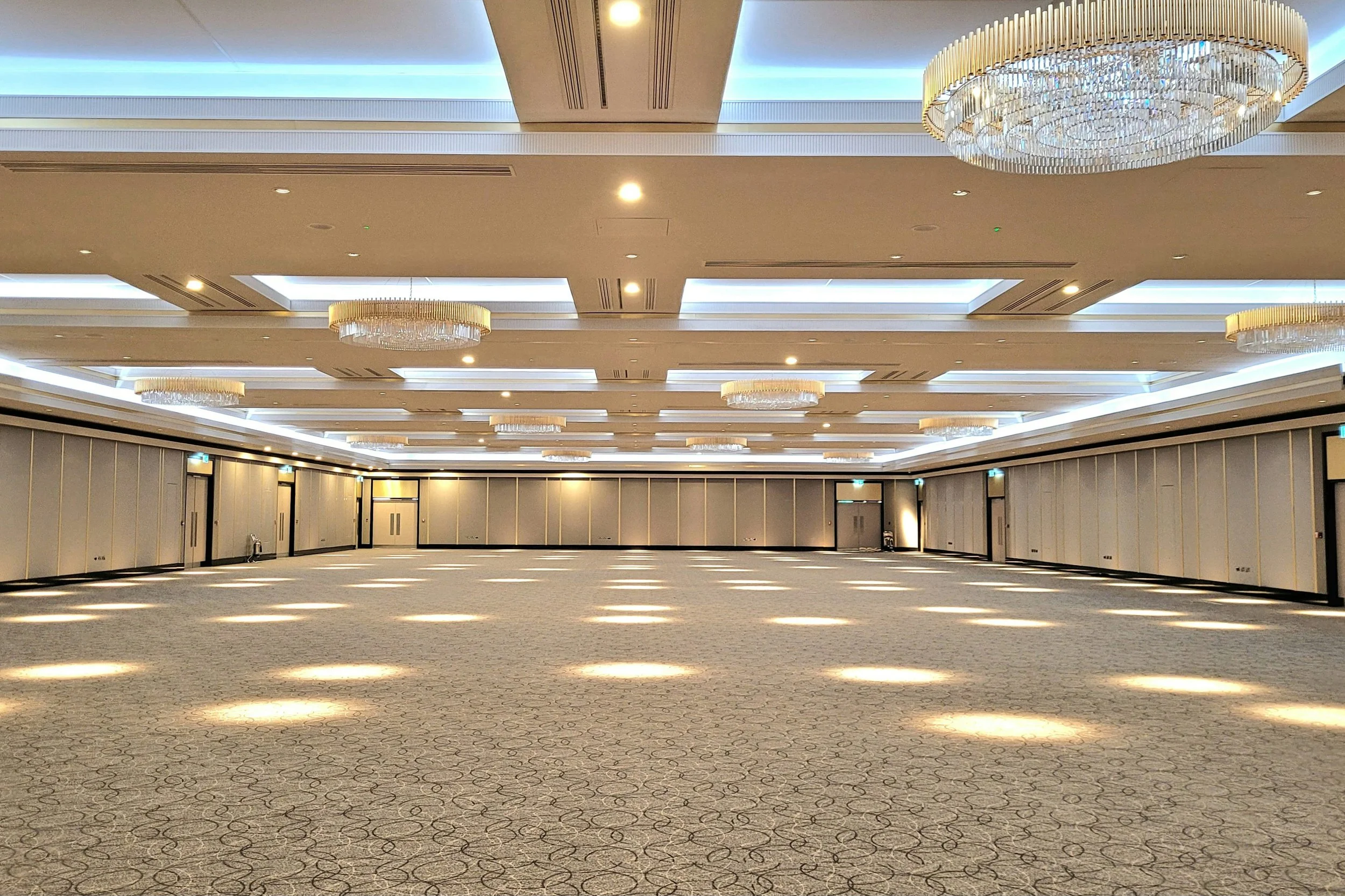 Large empty conference room with patterned carpet, beige walls, and ceiling-mounted rectangular light fixtures. Multiple elegant chandeliers hang from the ceiling, and there are doors along the walls.