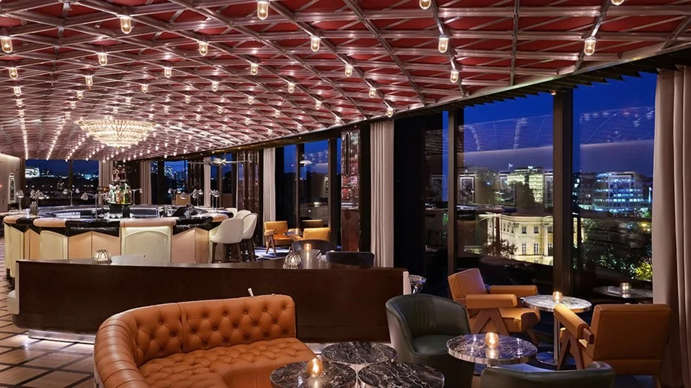 Interior of a luxurious bar or lounge at night with large windows, elegant seating, and a chandelier, city skyline visible outside.