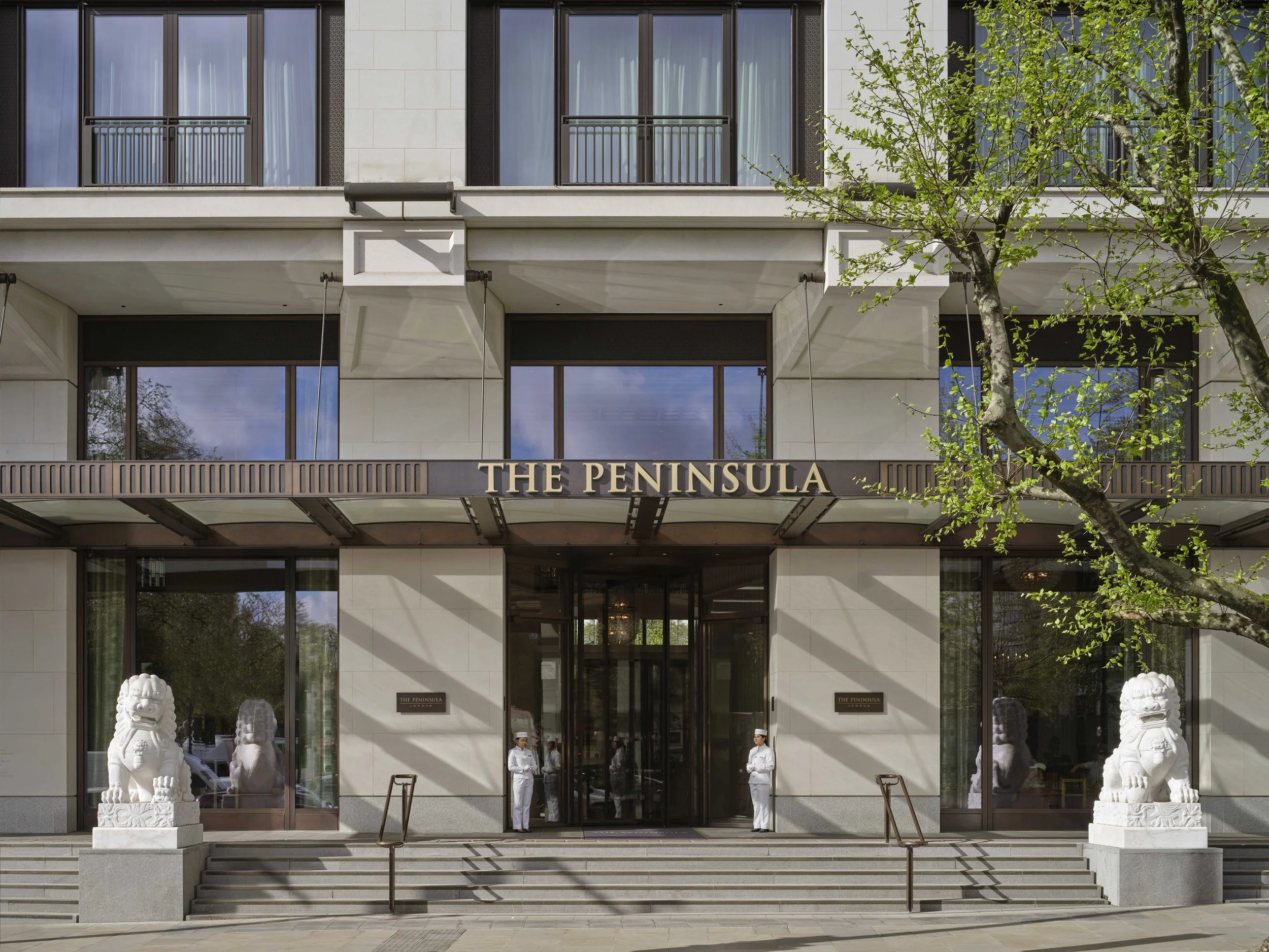 The entrance of The Peninsula hotel with white stone lion statues on either side of the steps, two staff members standing at the entrance, and a large glass door. The building has large glass windows, some with blue curtains, and a tree partially blo