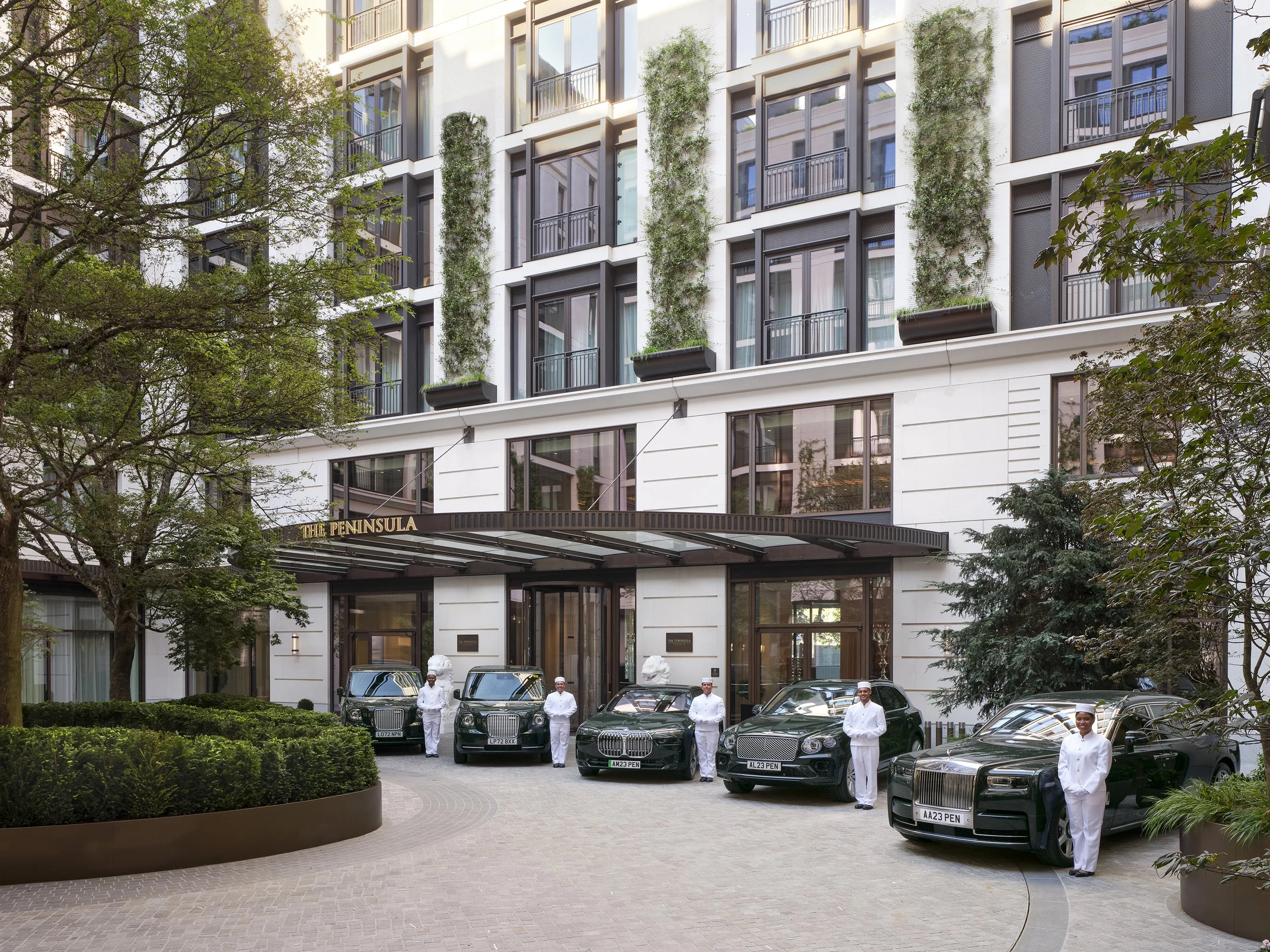 Luxury hotel entrance with a canopy, black cars parked in front, and staff standing by the vehicles. Modern building facade with balconies and greenery.