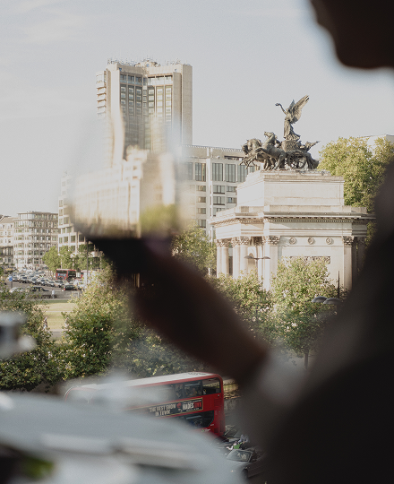View of a cityscape with a monument featuring a chariot with winged horses and a winged figure, background includes tall buildings, trees, and a red double-decker bus.