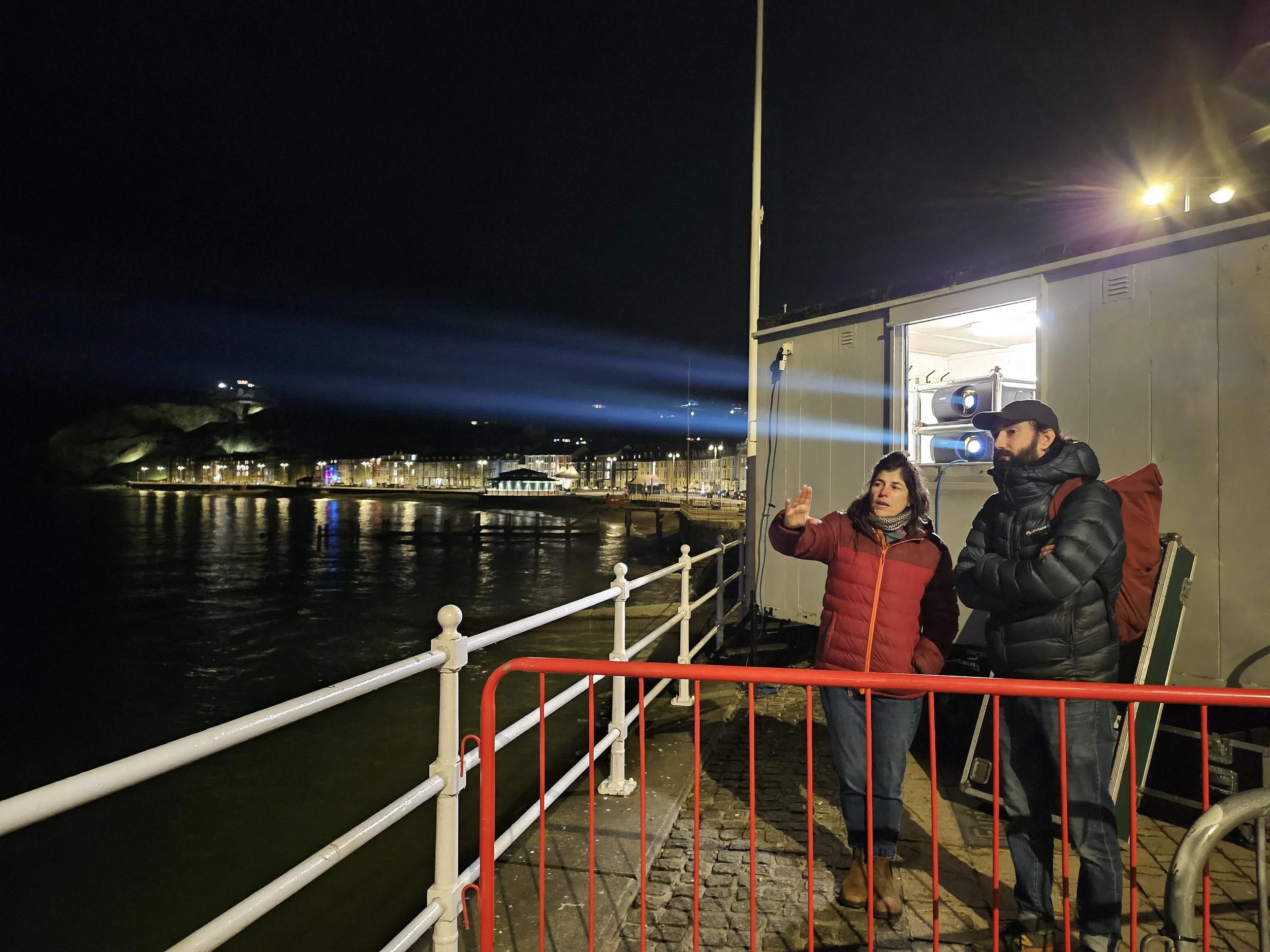 Two people standing on a dock at night, observing and discussing the nearby shoreline with illuminated buildings, a hill, and beams of blue light in the sky. One woman is gesturing while the man listens with arms crossed, both wearing winter jackets.