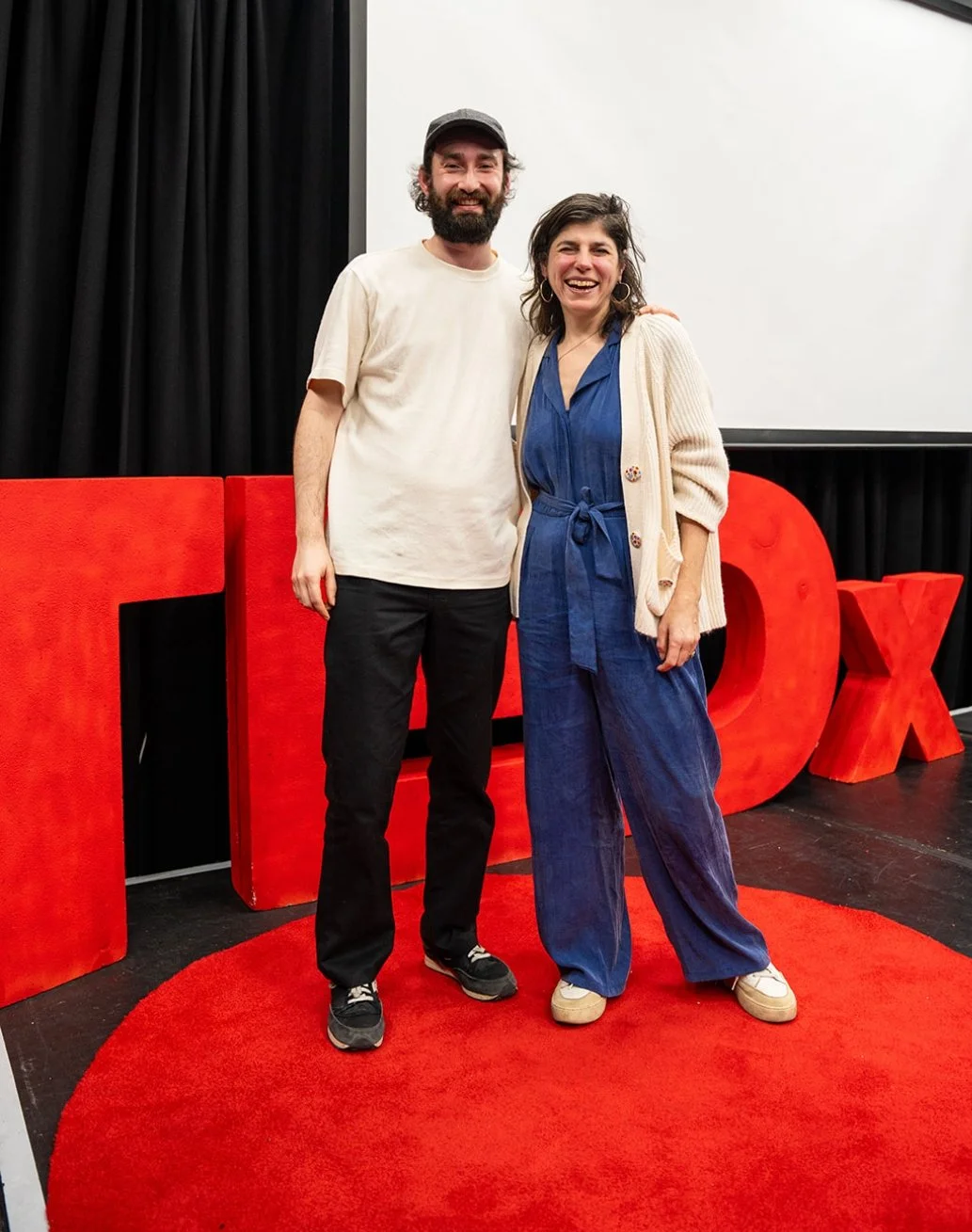 A man and a woman stand together on a red circular carpet, smiling at the camera. Behind them are large red TEDx letters and a black curtain, with a white screen to the right.