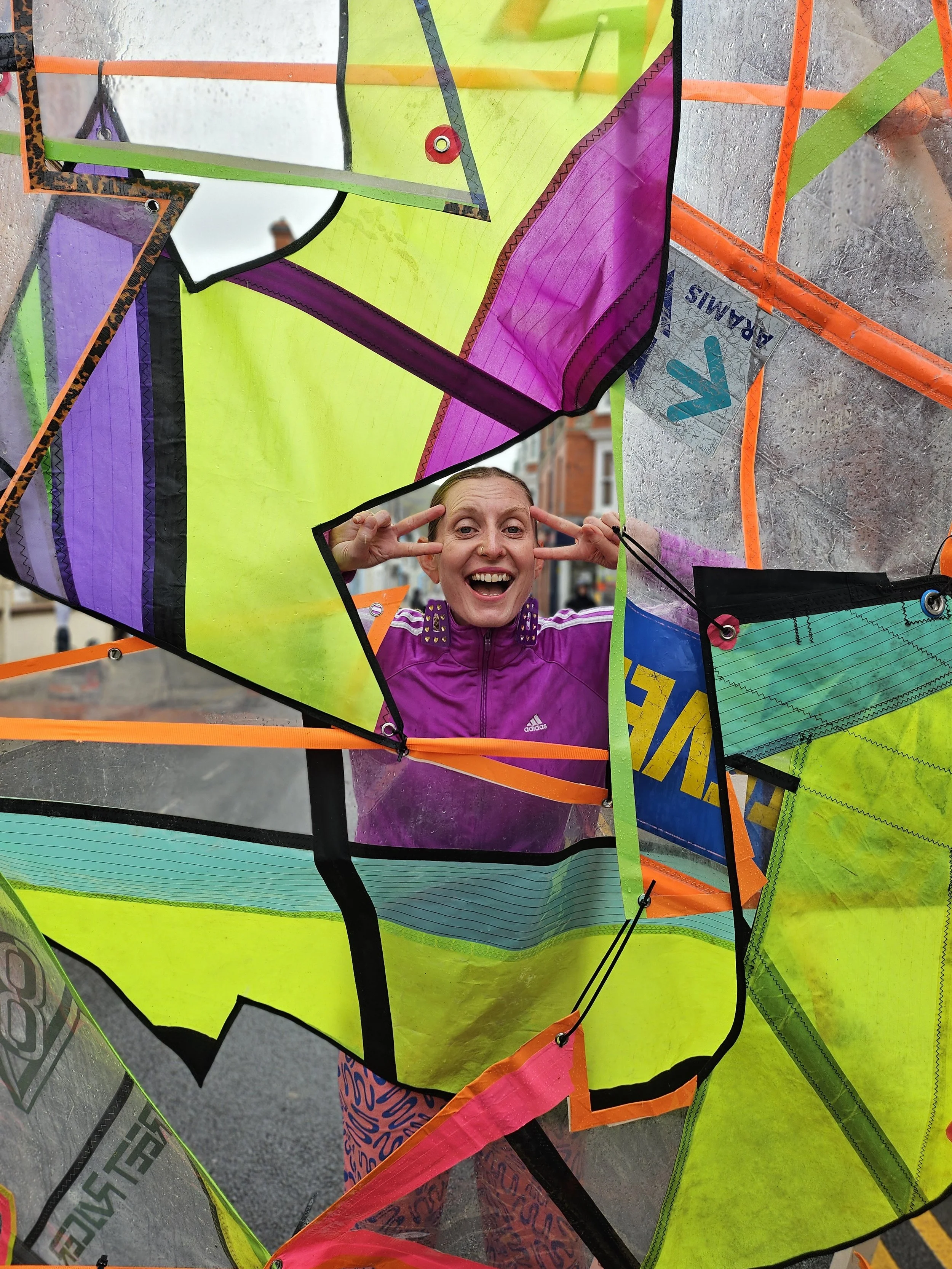 A woman in a purple jacket is smiling and making a peace sign with both hands while sticking her head through a hole in a colorful, geometric, translucent structure made of fabric and plastic.