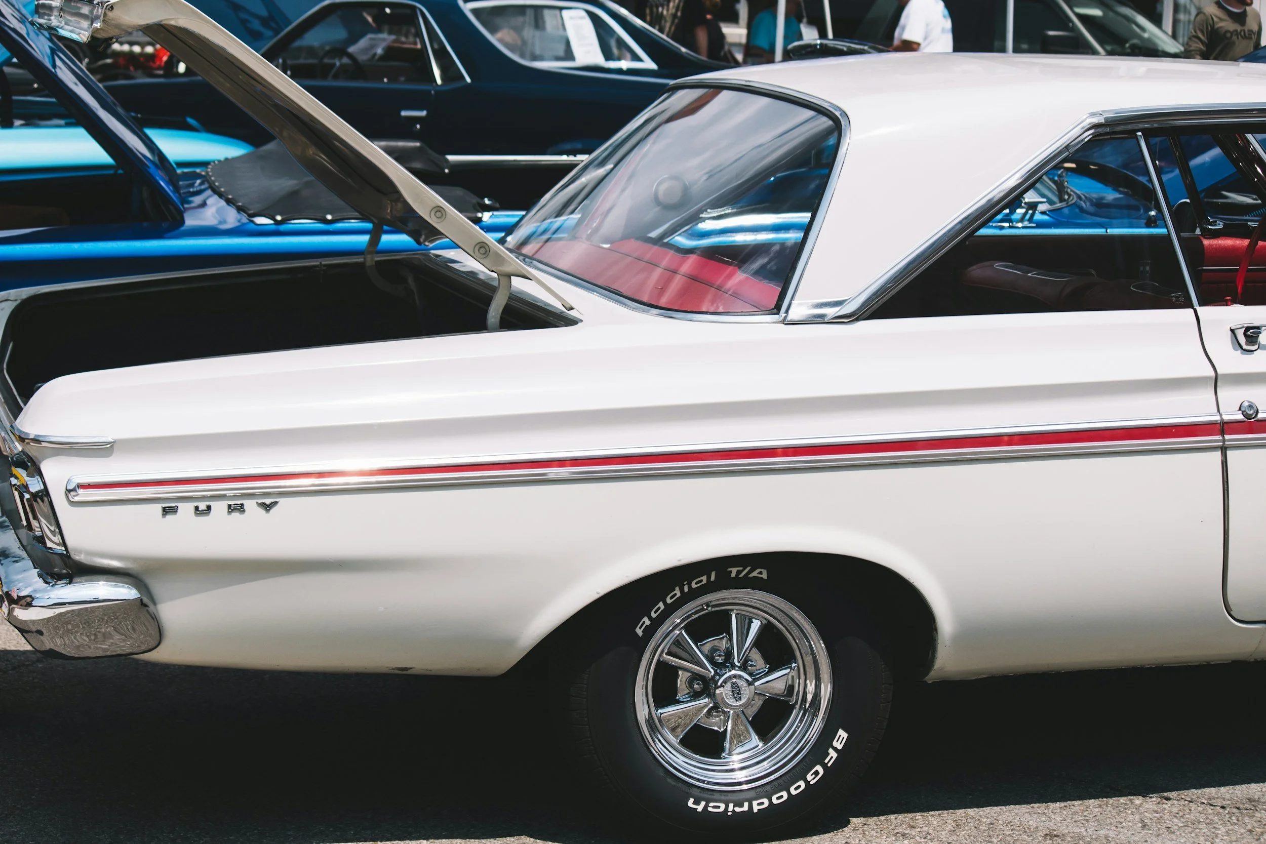 A white vintage muscle car with the trunk open at a car show, surrounded by other classic cars and people.
