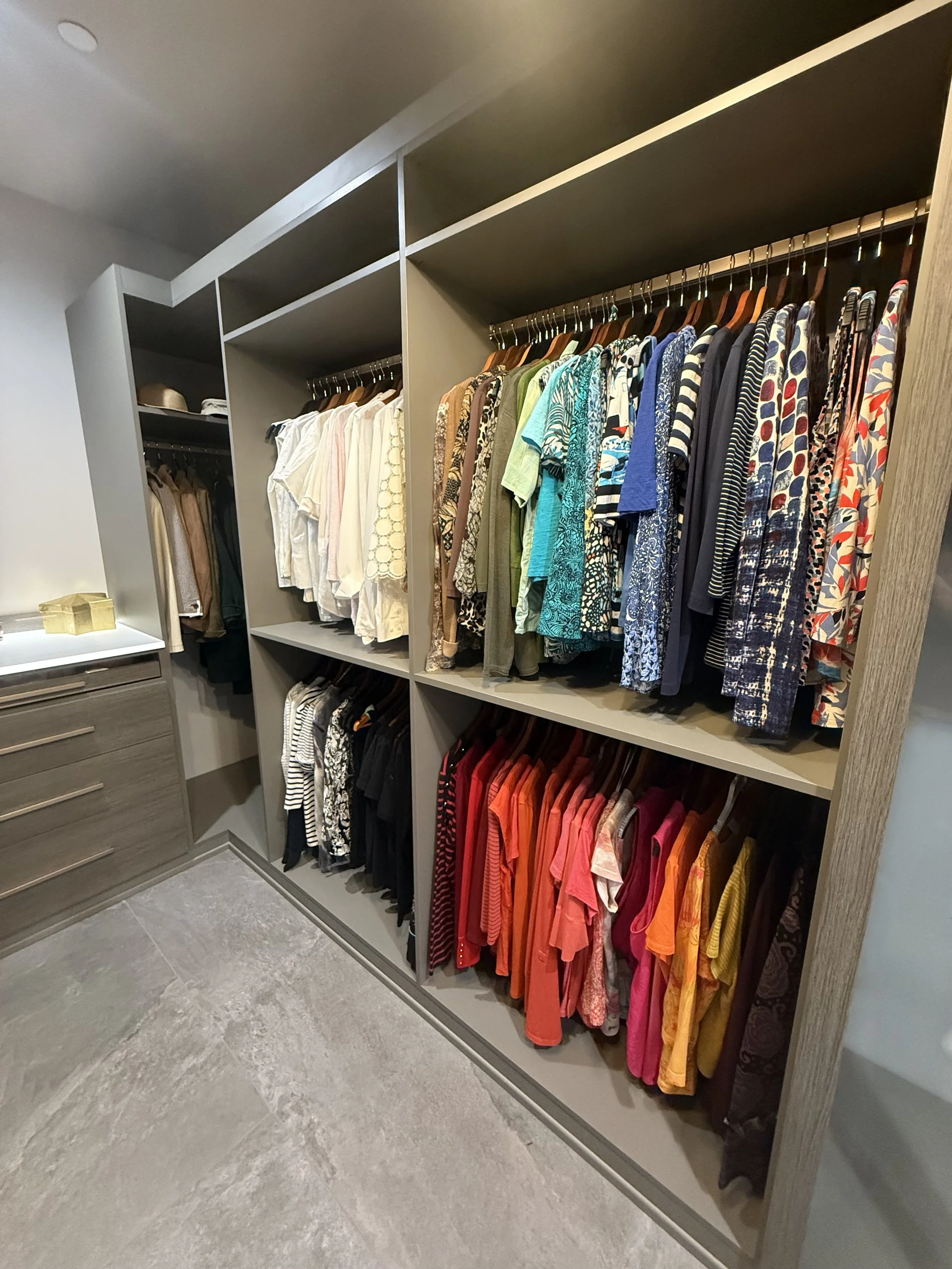 A closet with neatly organized clothing on multiple racks, including colorful shirts and blouses, with drawers on the left side.
