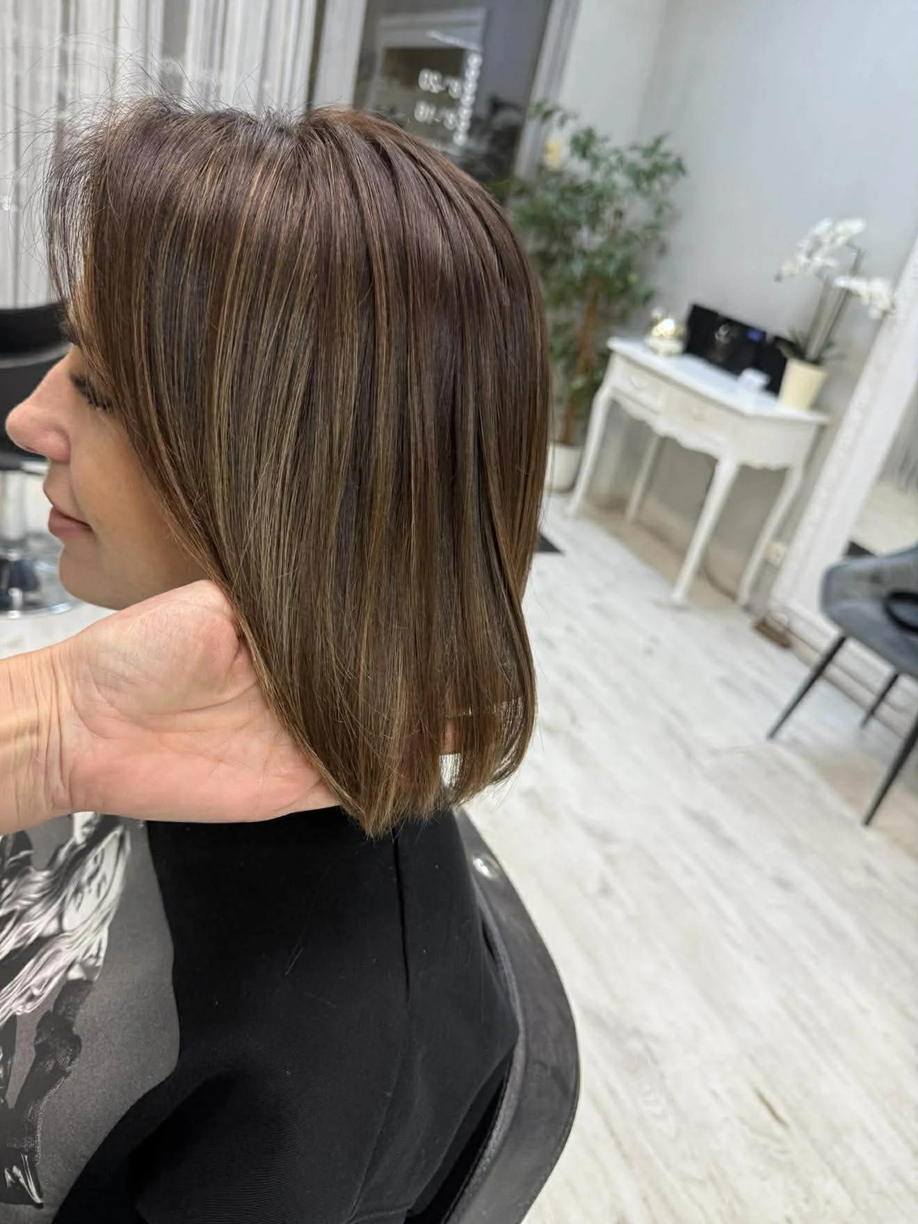 Moteris kirpėjo kėdėje: rudi plaukai iki pečių su šviesiomis sruogelėmis.