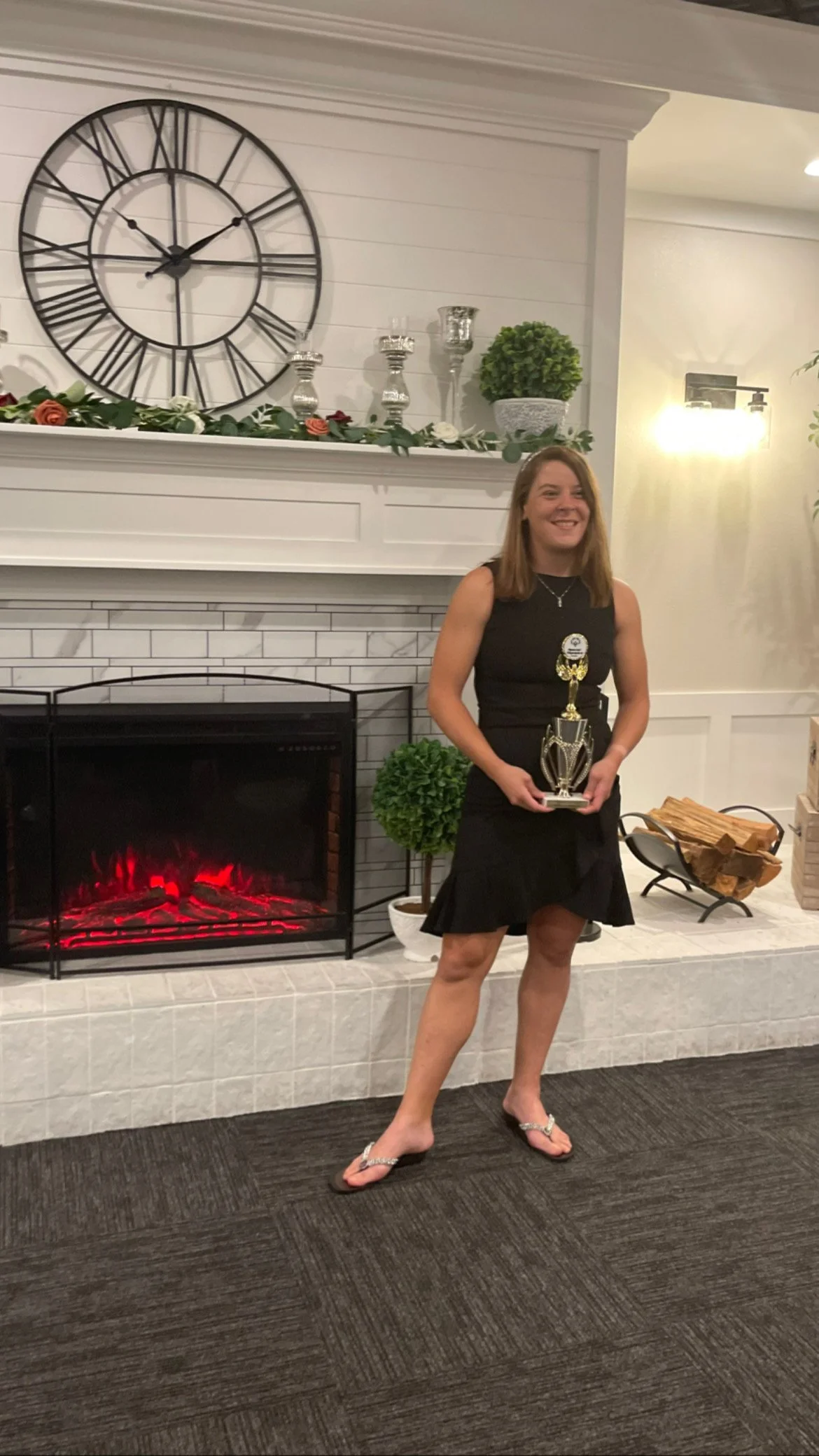 A woman in a black dress standing by a fireplace holding a trophy, smiling, and wearing flip-flops, with a large wall clock and decorative items on the mantel behind her.