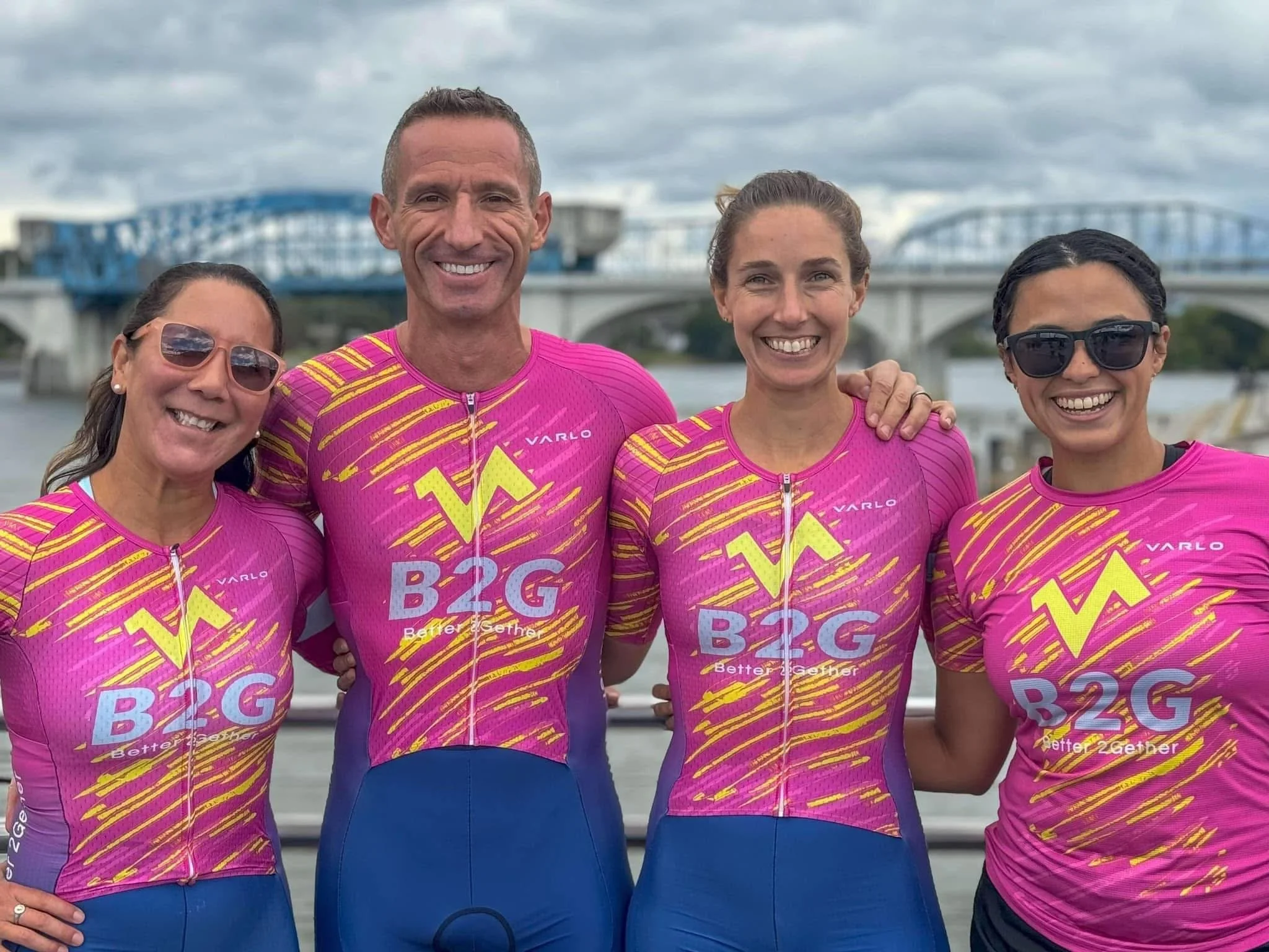 Four people smiling and posing together for a photo outdoors near water and a bridge, wearing matching pink and purple cycling jerseys with yellow and blue accents.