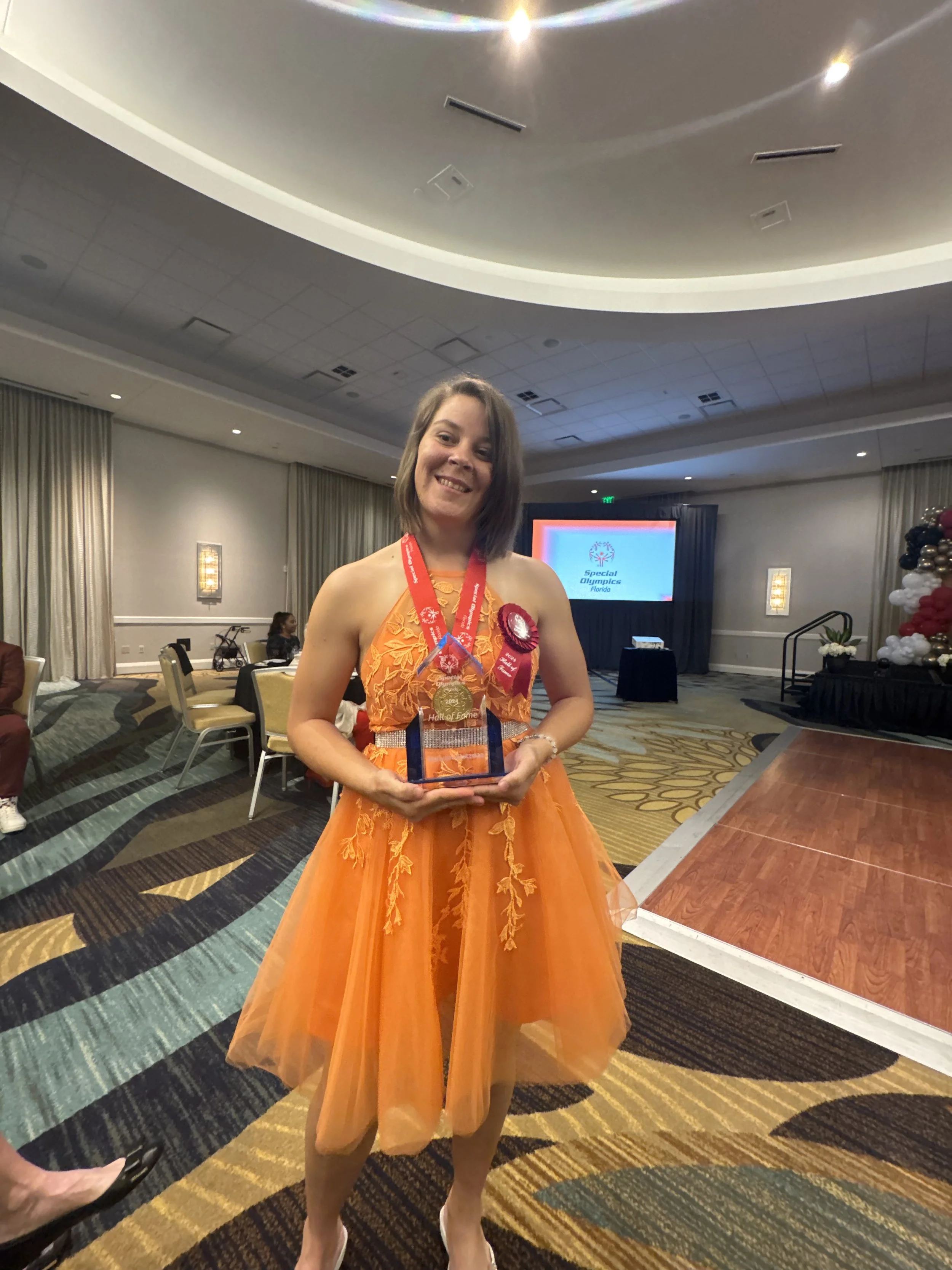 A woman in an orange dress holding a trophy, wearing medals and ribbons at an indoor awards event.