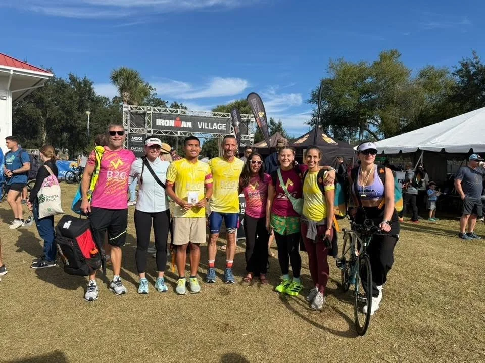 Group of people in athletic clothing at an outdoor event, with a banner reading 'IRONMAN VILLAGE' behind them, some holding medals, some with bicycles, under a blue sky with trees and tents in the background.