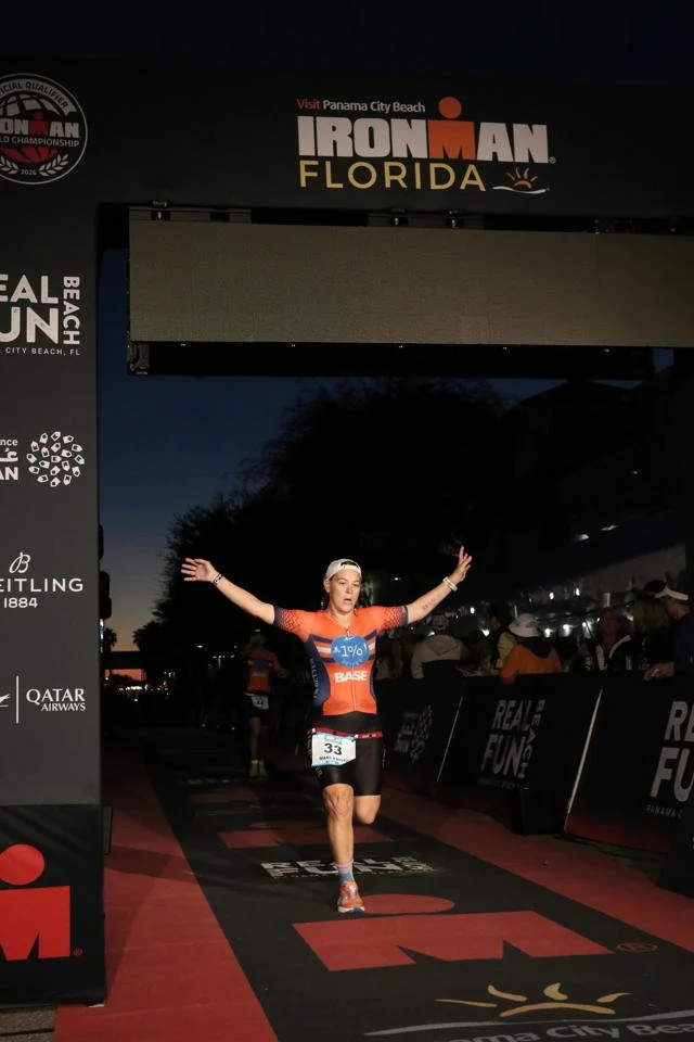 Marlynne Stutzman crossing the finish line of an Ironman race in Florida at dusk, wearing a medal around her neck, with her arms raised in victory, under a large IRONMAN Florida sign.