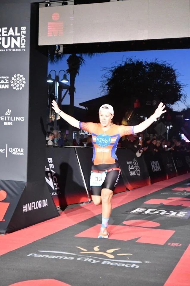 Female athlete crossing the finish line at a race event at Panama City Beach, celebrating with arms raised, wearing sports gear, with a crowd and event banners in the background.