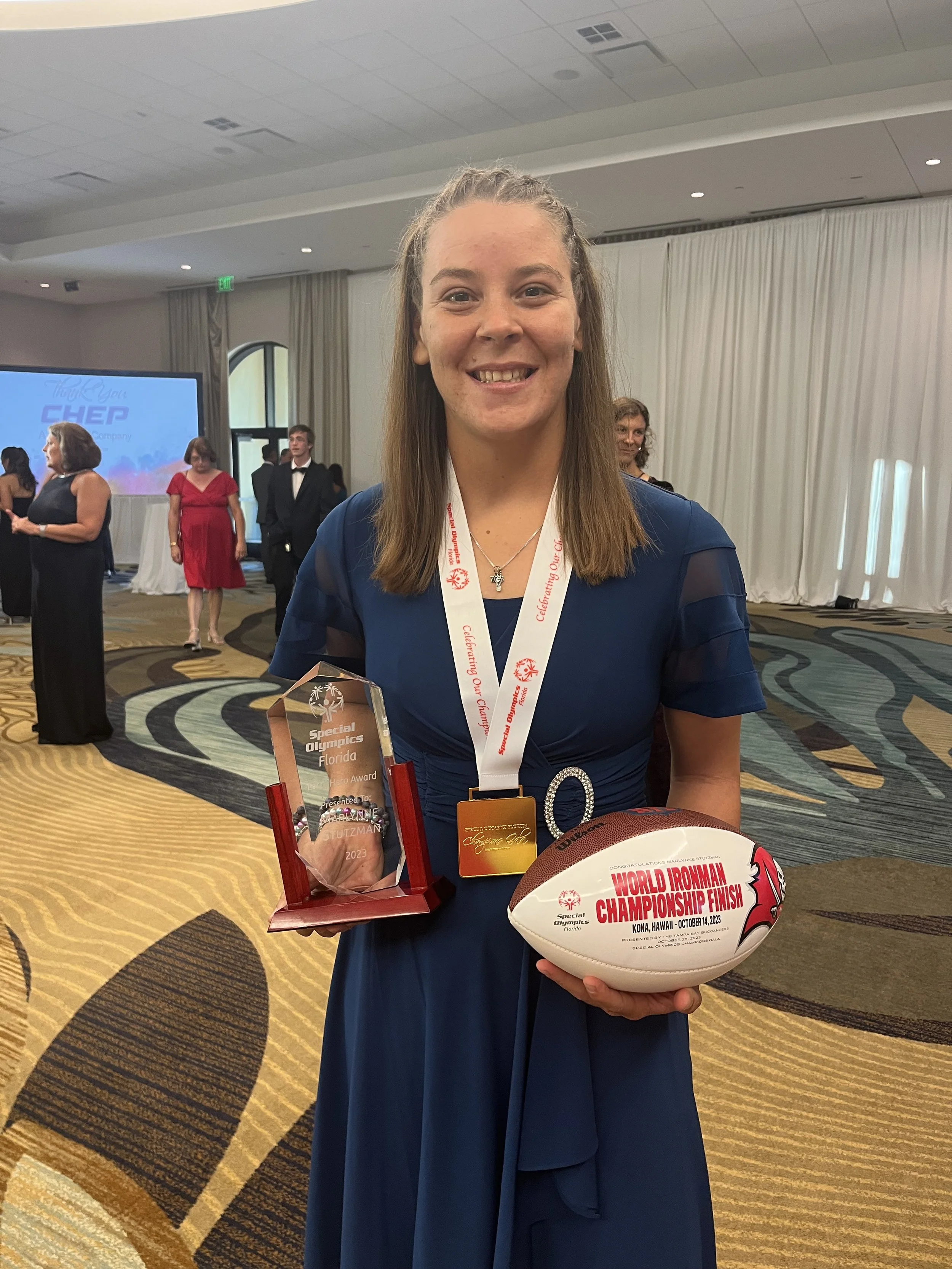 Young woman in a blue dress holding a Special Olympics Florida award, a World Ironman Championship finish football, and wearing a medal at an indoor event.