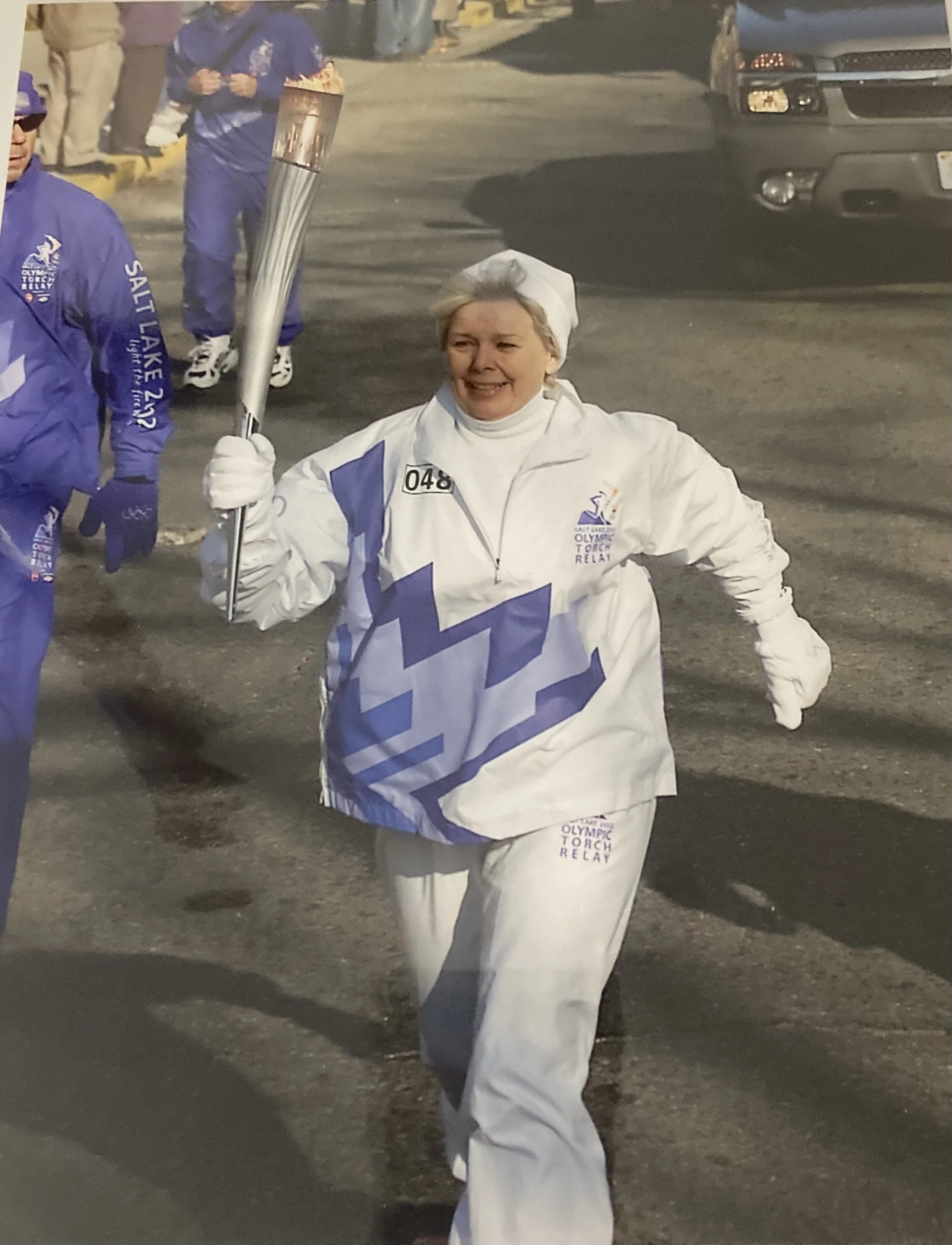 Woman running with a torch during the Salt Lake 2002 Winter Olympics torch relay, wearing a white jacket and pants, with others in similar attire behind her.